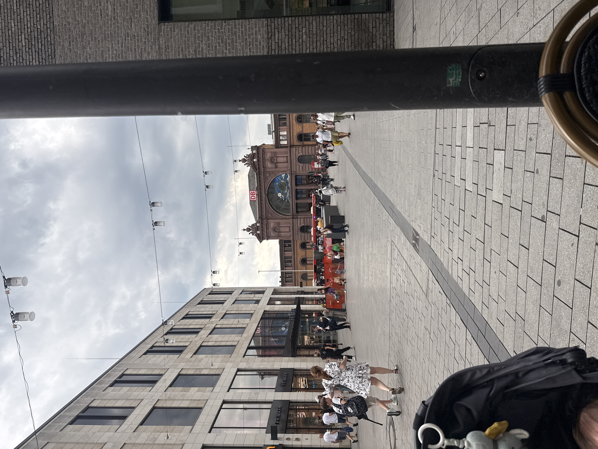 This is a vertically oriented, eye-level, full-shot photograph of a bustling city street scene in Bonn, Germany. The image is dominated by a large, dark pole in the foreground, partially obscuring the view. Beyond the pole, a wide pedestrian walkway paved with light grey stone bricks stretches into the distance.
On the left side of the walkway, a modern building with a light-colored stone facade and multiple dark-framed windows rises up. On the right side, in the mid-ground, is a grand, older building with a reddish-brown brick facade and arched windows, appearing to be a train station. A large clock face is visible within one of the arches. The building has architectural embellishments and a prominent "DB" logo, indicating it is a Deutsche Bahn (German Railways) station.
Numerous people are walking along the pedestrian area, some singly, some in small groups. They are dressed in casual summer clothing. The activity appears to be ordinary street life and transit, with people moving in various directions.
In the background, a red bus is visible. Overhead, a network of wires with small lights strung across them suggests it might be evening or an overcast day, though the sky is a cloudy grey.
Notable details include the "Primark" text visible on the modern building on the left, indicating a retail presence. The overall mood is one of urban activity and movement. The lighting suggests it could be late afternoon or early evening.