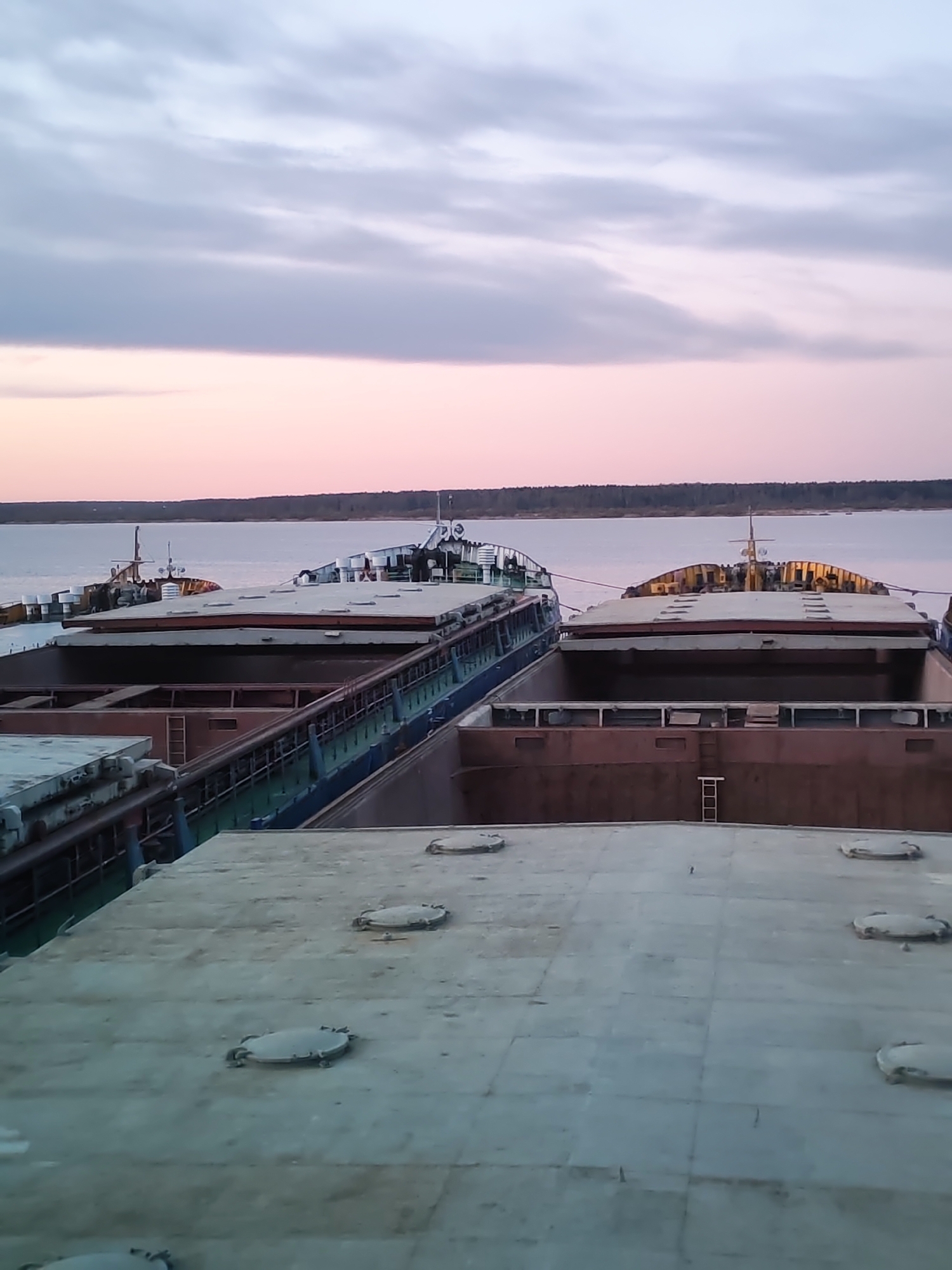 The image depicts a view from the top of a barge in Rybinsk, Russia. Several barges are moored side by side on a river, with visible hatches on their decks. The background shows a river bordered by a distant treeline and a sky with a gradient of pink and gray clouds at dusk. No people are visible in the image.