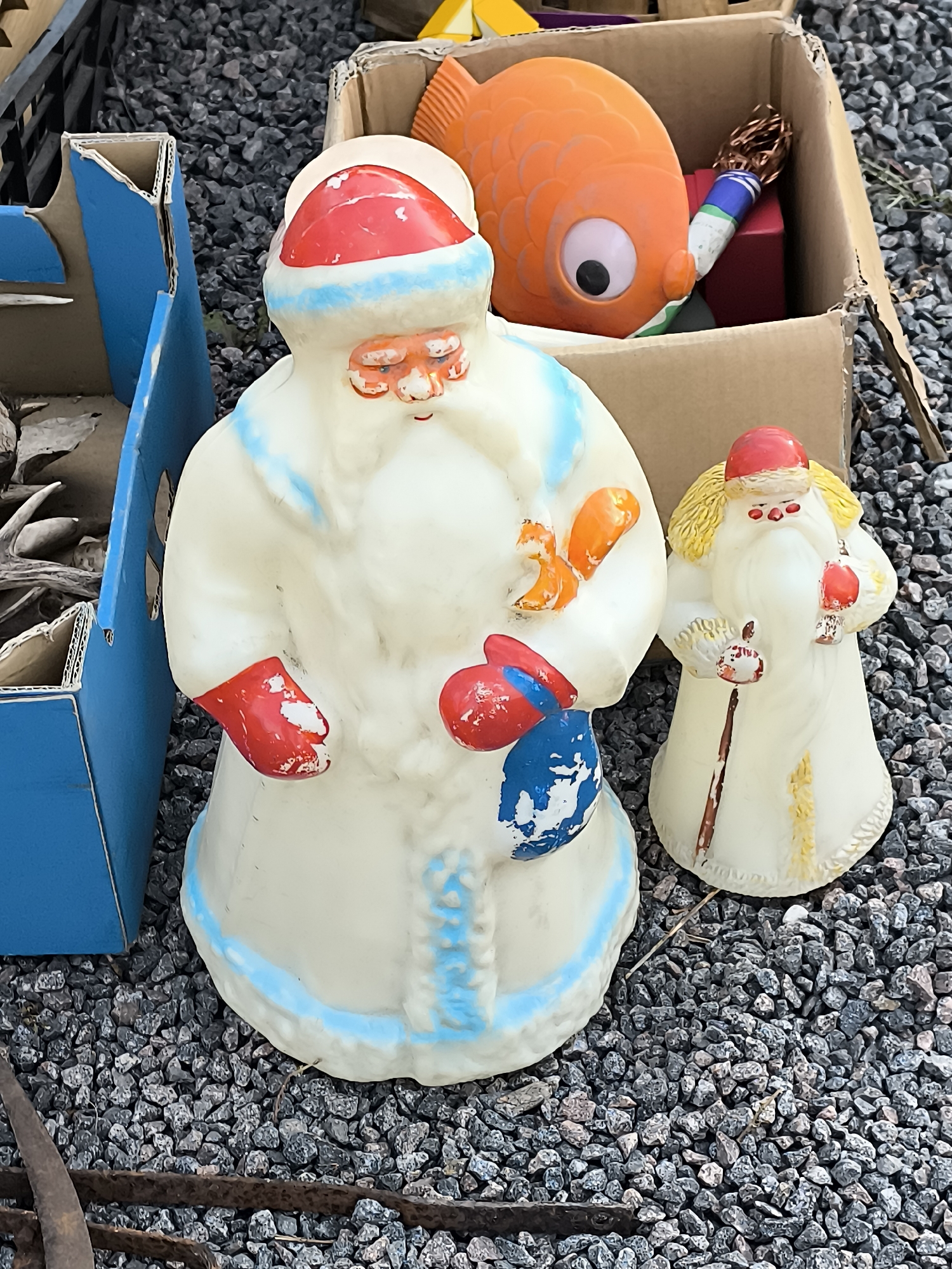 This is a medium-shot, eye-level photograph of two vintage, blow-mold Christmas decorations, likely depicting Santa Claus figures, placed on a gravel surface. The figures are made of translucent plastic with painted details.
The larger figure on the left is predominantly white with red and blue accents. It wears a red hat with a blue trim, a white coat with blue trim, red mittens, and holds a blue bag. A small, orange squirrel figure is attached to its right hand. The paint on the mittens and bag shows signs of wear and chipping.
The smaller figure on the right is also white with red and yellow accents. It has a red hat, a white coat with yellow trim, and holds a staff with a red-tipped ornament. The paint on this figure also appears worn in places.
To the left of the figures is a portion of a blue cardboard box, open at the top, revealing some brown cardboard edges and what appears to be dried foliage or branches. To the right and behind the figures is a cardboard box containing a large, orange, fish-shaped toy with a prominent eye, and a cylindrical object with colorful bands and a copper-colored brush-like end. The background consists of more gravel and some indistinct darker objects. The lighting suggests an outdoor setting, possibly during daylight hours.
The image does not contain any identifiable landmarks, signs, or contextual clues to place it specifically within Klaipėda, Lithuania.