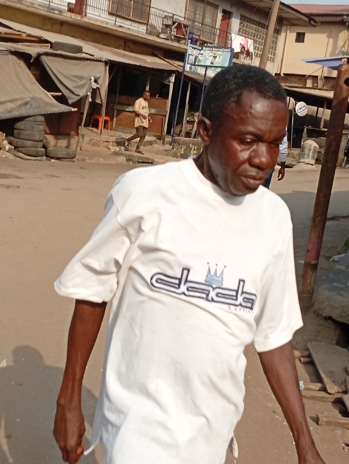 An older man in a white "dada" t-shirt walks outdoors, with a street scene behind him. Buildings and shops line the background. A younger man in a light-colored shirt is walking in the background. The setting appears to be a bustling urban area, possibly a marketplace or commercial district. The lighting suggests it is daytime.