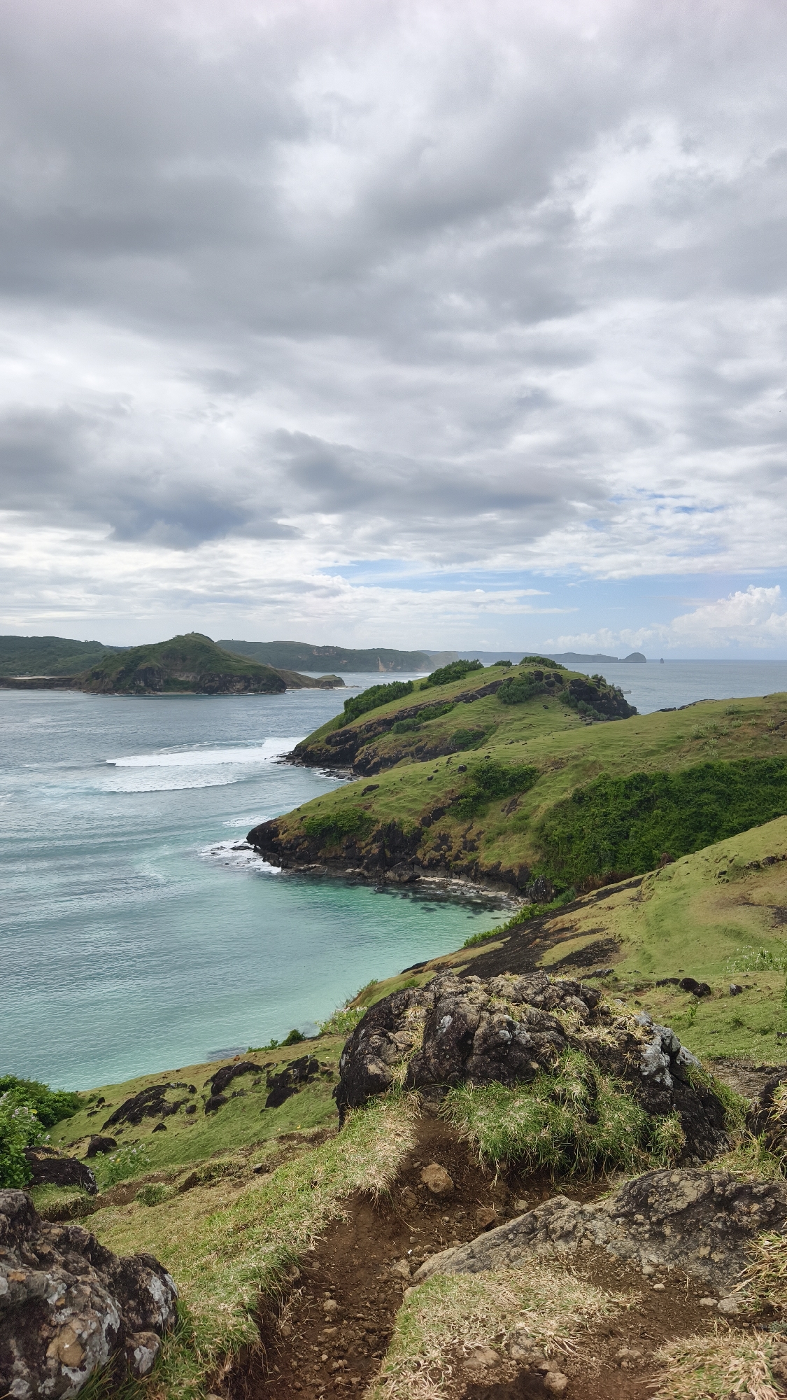 This image presents a sweeping coastal landscape view, likely captured from a high vantage point in Kuta, Indonesia. The scene is dominated by lush green hills, turquoise ocean waters, and an overcast sky.
In the immediate foreground, a rugged, earthy path winds downwards through patches of green grass and dark, moss-covered rocks. This area suggests a natural, somewhat unmaintained trail or observation point.
The midground showcases a series of prominent, verdant headlands that extend into the sea. These hills are covered in dense green grass, contrasting with the dark rock formations along their edges where they meet the water. The ocean itself displays a gradient of colors, from clear, light turquoise in the shallower coves to deeper blues further out. White, frothy waves can be seen breaking as they approach the shorelines, particularly along the left side of the frame.
In the background, more distant landmasses or islands are visible, stretching across the horizon, appearing somewhat hazy.
The sky is largely covered with thick, grey clouds, indicating an overcast day. However, patches of lighter sky and faint blue are visible towards the horizon on the right, suggesting indirect daylight. The overall lighting indicates it is daytime, possibly late morning or early afternoon.
No people, man-made structures, or visible text are present in the image, emphasizing the raw, natural beauty and tranquility of the coastal environment.