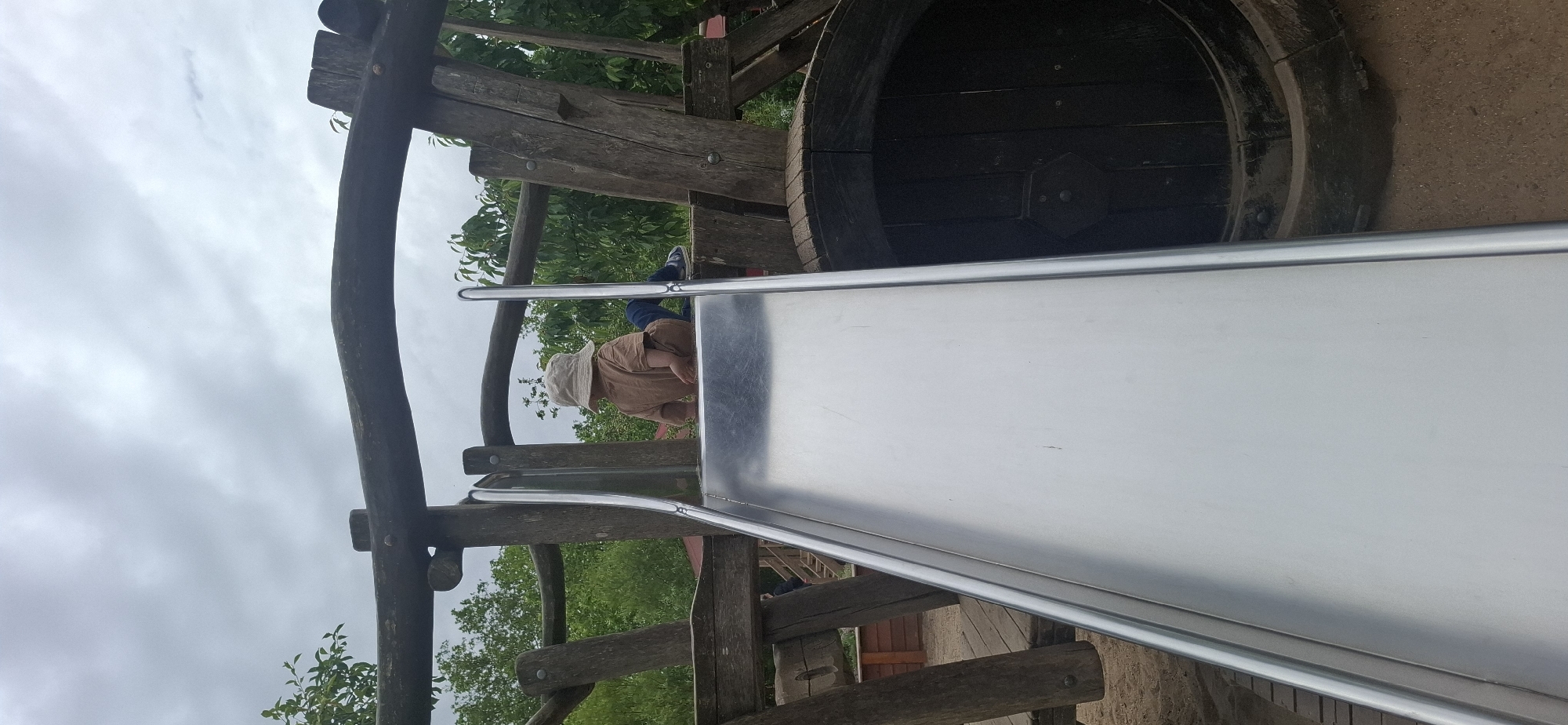A child wearing a tan hat and shirt sits on a silver slide at a playground. The child is facing away from the camera, looking down the slide. The playground structure is made of wood and metal, with a large barrel-like structure to the right of the slide. The background shows trees and a cloudy sky. The playground is likely located at the Naturbad in Hürth, Germany, a popular recreational area known for its natural swimming pond and adventure playground.