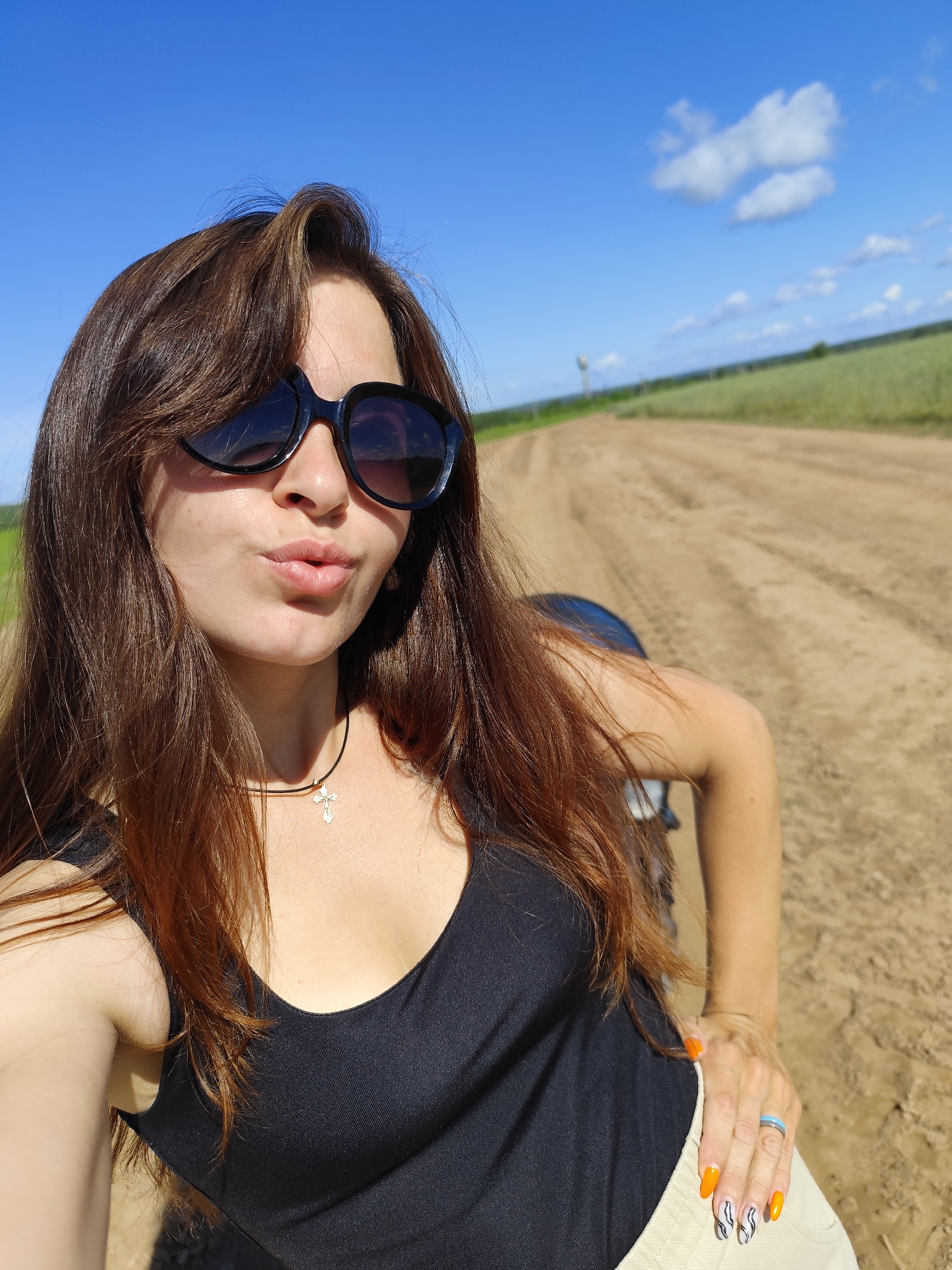 In the city of Фоки, Russia, a young woman with long, brown hair and black sunglasses is taking a selfie outdoors. She is wearing a black tank top, tan shorts, and a cross necklace. Her lips are pursed, and she has a blue ring on her right hand with painted fingernails. She is standing on a dirt road with a field and blue sky in the background. The overall scene suggests a casual outdoor activity on a sunny day.