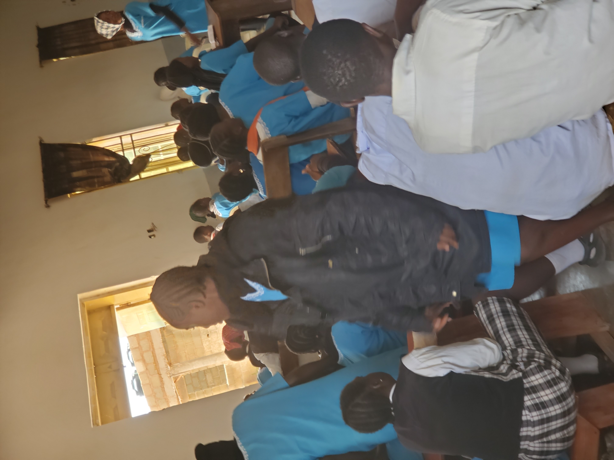 The media file shows a group of people, predominantly young individuals, in what appears to be a classroom setting in Kuru Karama, Nigeria. The subjects are seated at wooden desks, some with their heads bowed, suggesting they might be engaged in learning, a prayer, or some form of instruction. The majority of the individuals are wearing blue uniforms, possibly indicating a school or a specific organization. One person in the foreground is wearing a black jacket over a blue shirt, and another person is in a white shirt and light-colored trousers. The room has large windows with dark curtains, and natural light is entering from them, suggesting it is daytime. The walls are a light color, and the overall atmosphere seems calm and focused. There is a door visible in the background, partially open, offering a glimpse of the outside environment. The exact activity is not definitively clear, but the posture of the individuals suggests a period of concentration or reflection. There is no visible text in the image.