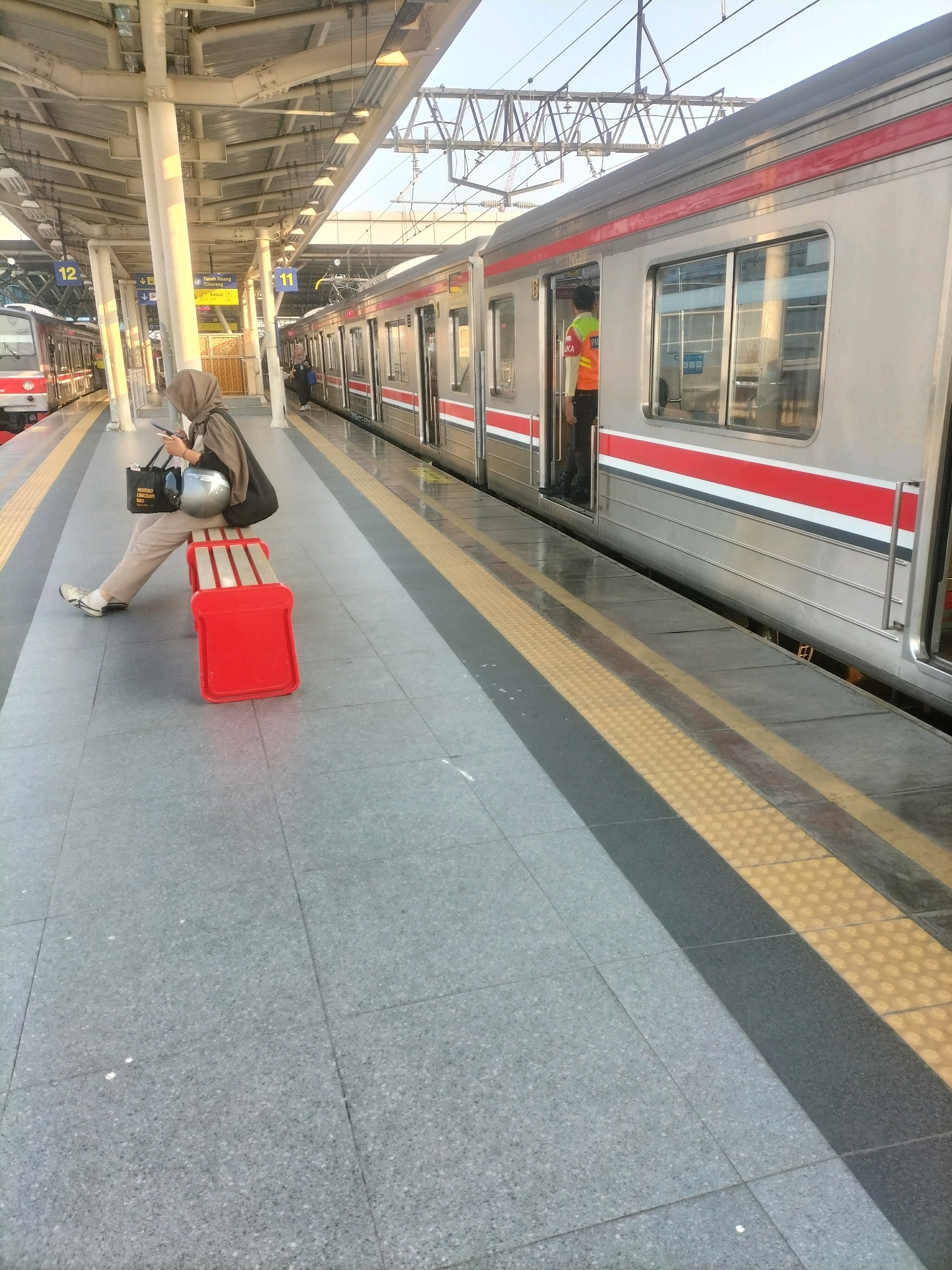 The image depicts a train station platform with two trains present. A woman wearing a hijab is seated on a bright red bench to the left of the frame, holding a black tote bag and a silver helmet. She is looking at her phone. The platform has a textured surface with yellow and black safety lines. A train car, with silver siding and red and white stripes, is stopped at the platform on the right. A person in a bright orange vest is standing in the open doorway of this train car. Overhead, there are station signs indicating platform numbers 11 and 12, and the name "Tanah Abang Citayam". The overall environment suggests a sunny day at a public transportation hub.