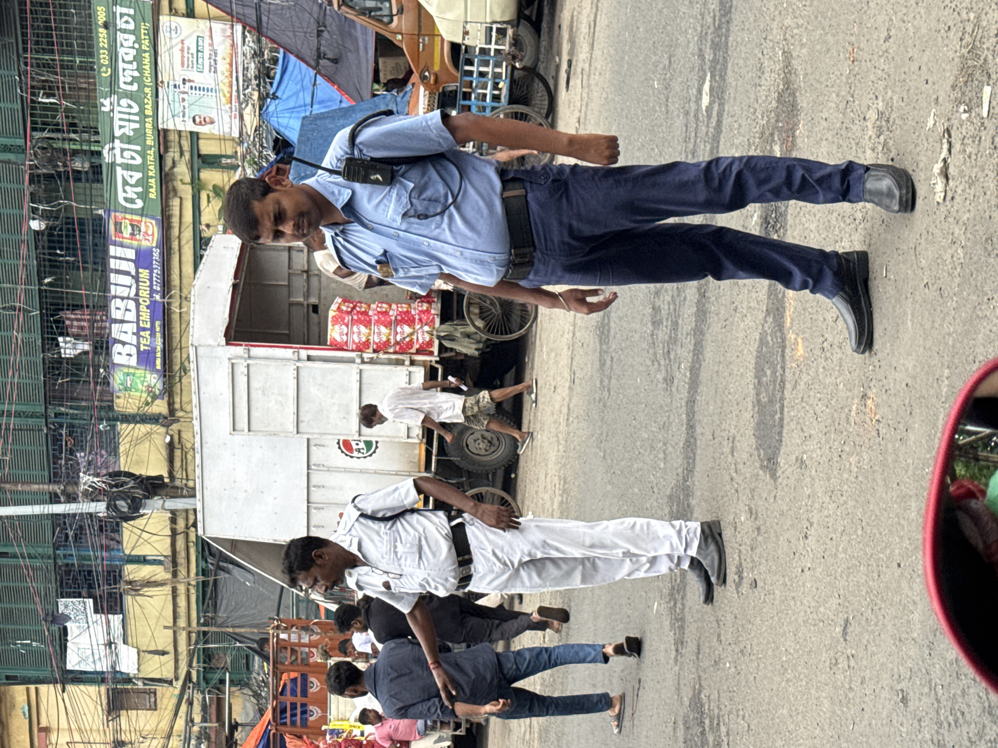 The image shows a street scene in Kolkata, India, likely in the Burrabazar area, a busy commercial district known for its trading and markets. Two traffic police officers are present. One officer, in the foreground, is wearing a light blue uniform, dark blue pants, and black shoes, with a radio on his shoulder. The other officer, to the left and slightly behind, is dressed in a white uniform and white pants with black shoes, and appears to be gesturing with his right hand. Several other individuals, dressed in casual clothing, are visible in the background, along with a large white truck, a partial view of a brown truck, and various market stalls and buildings. The environment is urban and congested, with overhead wires and signage typical of a commercial street in Kolkata.