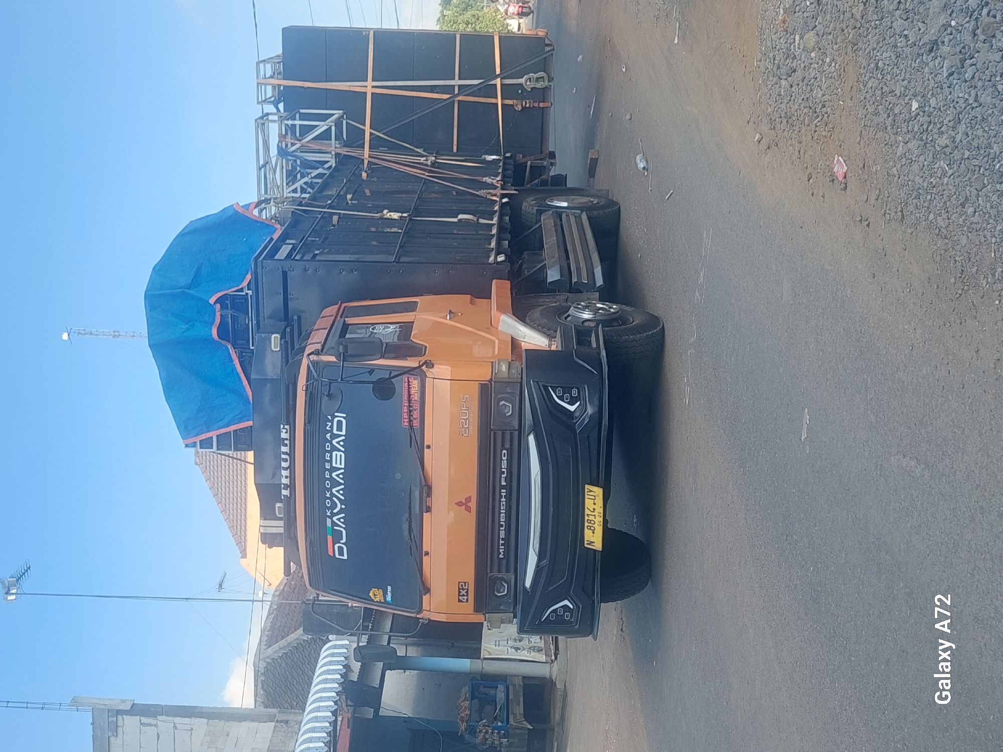 The image shows an orange Mitsubishi Fuso truck parked on a paved road in what appears to be a residential or semi-commercial area. The truck is modified and equipped with a large sound system, with speakers stacked on its rear. A blue tarp covers some of the equipment on top of the truck. The truck has the text "DJAYAAABADI" and "KOKOPERDANA" on its windshield, along with a "THULE" logo. A license plate with the number "N 8814 UY" is visible on the front. Buildings with tiled roofs and what looks like a small shop with awnings are present on the left side of the image. A utility pole with antennas is visible in the background. The road is lined with gravel and debris. The sky is clear and blue. The general environment suggests a daytime scene in a location that might be part of Sengguruh, Indonesia, given the context provided. There are no people clearly visible in the foreground or engaging in any specific action.
