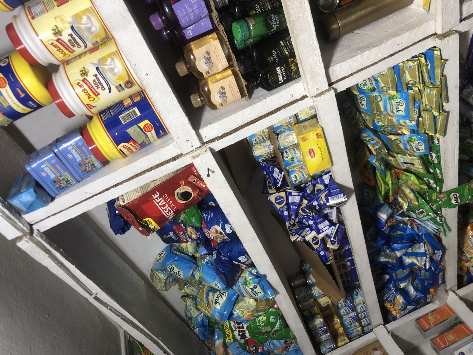 The image shows a cluttered shelf in a small store in Ado, Nigeria. The shelves are filled with various grocery items, including packets of milk powder, tea, and instant coffee. Some of the prominent brands visible are "Peak" milk, "Nescafe Classic" coffee, and "Lipton Yellow Label" tea. There are also several bottles of what appear to be alcoholic beverages, some with labels such as "Longstack Bitters" and "Kolaq Bitters." The items are arranged in a somewhat disorganized manner, with many packages piled on top of each other. The lighting is dim, suggesting an indoor setting, likely a small shop or a pantry. The overall impression is one of a well-stocked but cramped retail space.