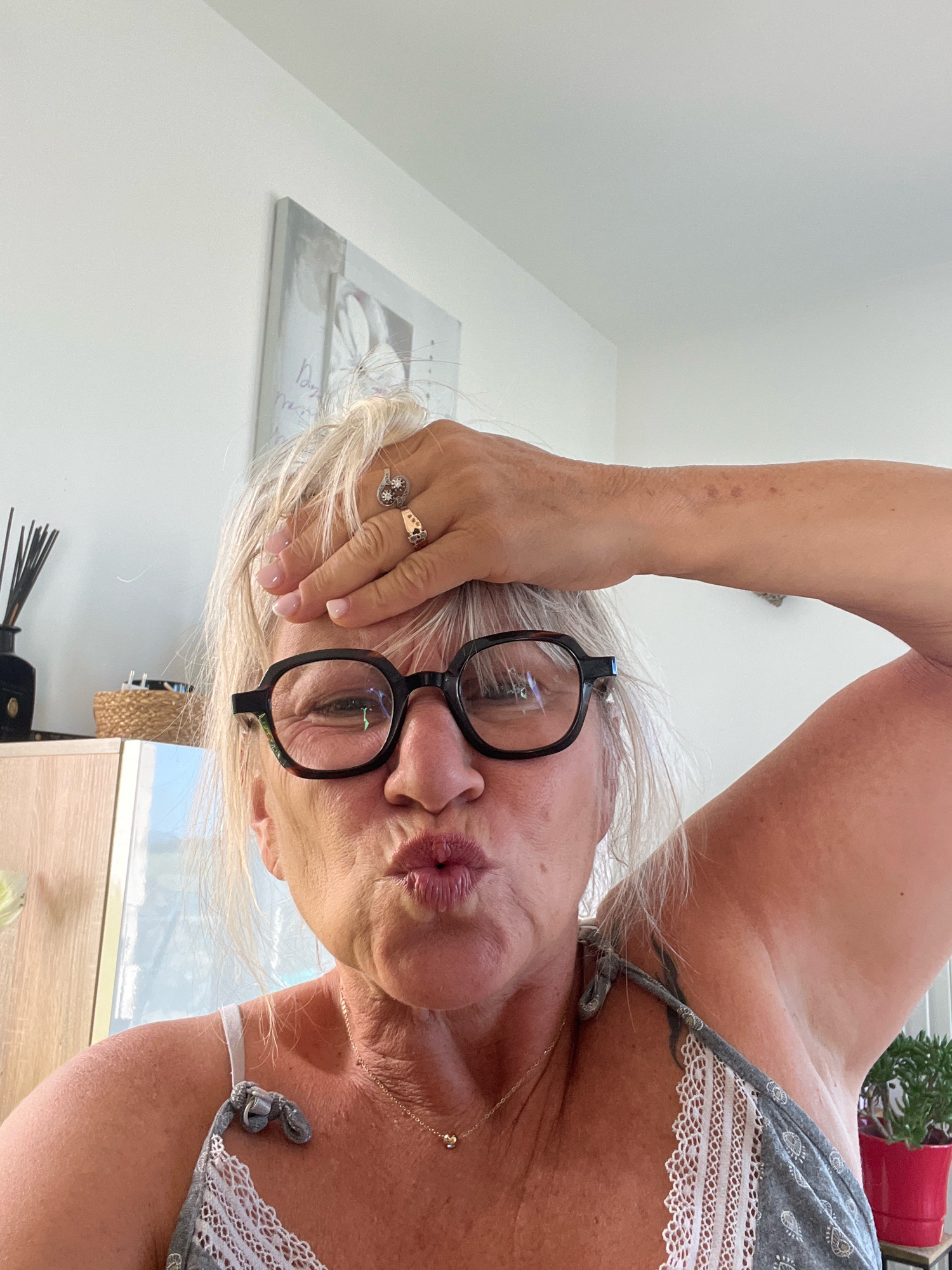 A close-up, eye-level selfie shows a Caucasian woman with blonde hair and glasses puckering her lips while holding her right hand to her forehead. She is wearing a grey and white patterned top with lace trim and a delicate gold necklace. Two rings are visible on her right hand. In the background, a white wall with a framed artwork is visible, along with a cabinet and a potted plant. The scene appears to be indoors, likely in a residential setting. There is no indication of the location being in Chambœuf, France.