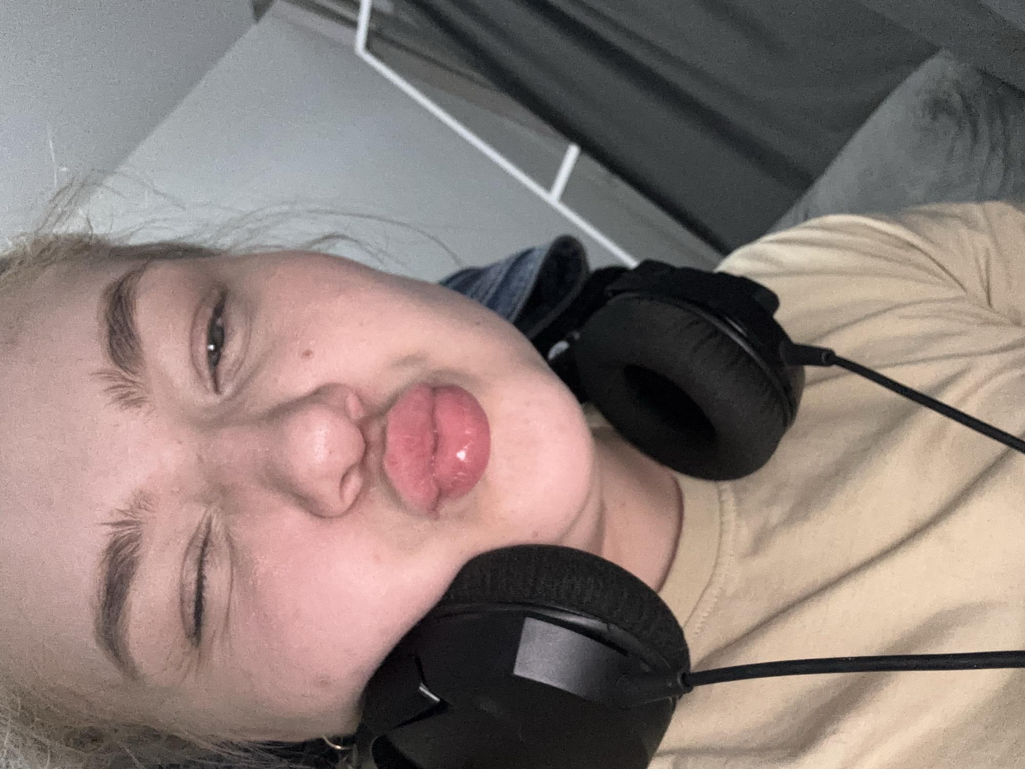 A person with fair skin is photographed close-up with their face taking up most of the frame. The person's eyes are closed, one eye appears to be winking, and their lips are pursed into a kiss. Black over-ear headphones are draped around the person's neck. The person is wearing a light tan t-shirt with a dark blue denim collar visible at the neck. The background includes a white-painted metal frame and a dark grey textured fabric or curtain.
There is no information to indicate the location is Chaikovsky, Russia.