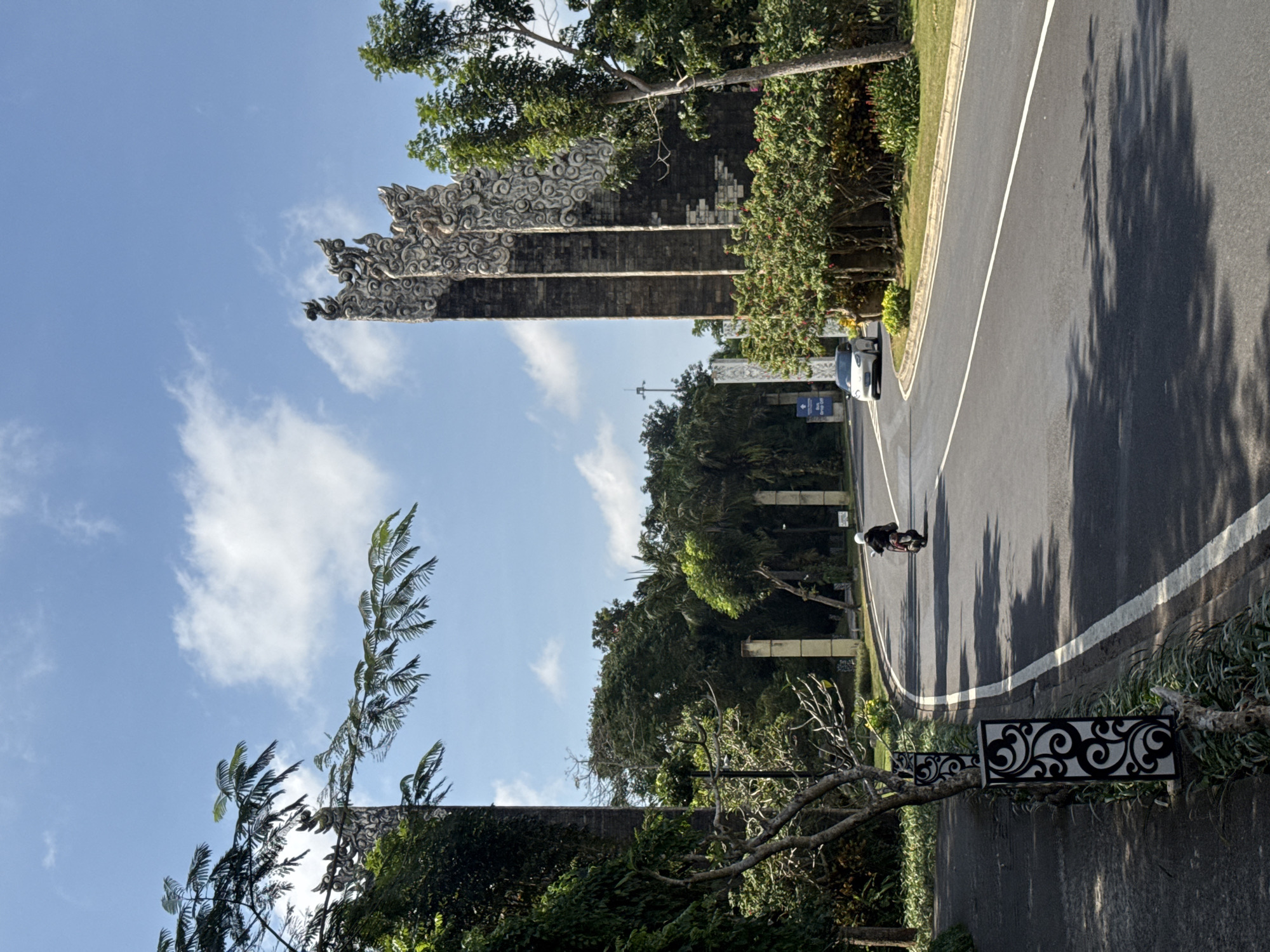The image shows a sunny day on a road in Kuta Selatan, Indonesia, with lush green trees and ornate stone gates framing the scene. A person on a scooter rides away from the camera on the right side of the asphalt road, which is marked with a white line. A white car is visible in the distance on the right, and a white sign is visible on the left. The stone gates are decorated with detailed carvings and appear to be part of an entrance to a complex or compound. The sky is blue with scattered white clouds.