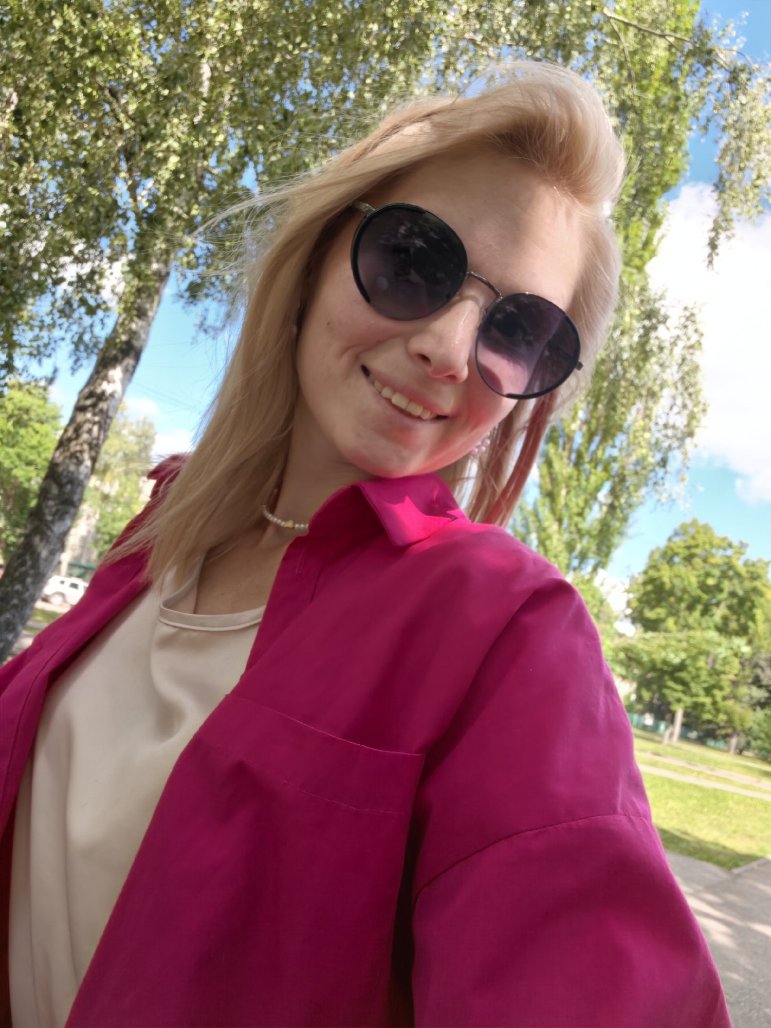A blonde woman, wearing sunglasses, a pearl necklace, a cream-colored top, and a vibrant pink shirt, smiles at the camera. She is outdoors, with trees and a blue sky visible in the background. The image appears to have been taken during the daytime, with natural lighting. The composition suggests a selfie, with the woman positioned in the foreground and the environment in the background. No specific landmarks of Sterlitamak, Russia, are identifiable in the image.