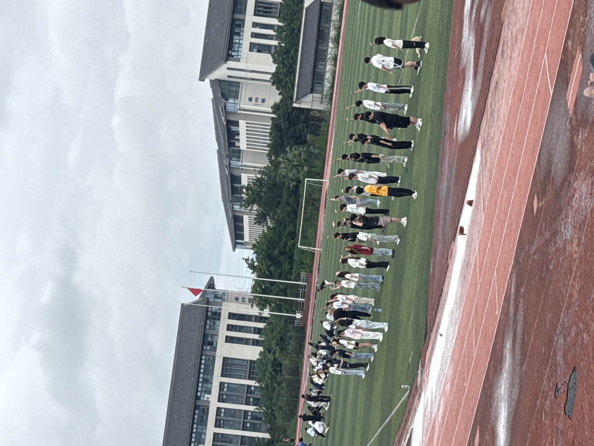 An outdoor, overcast daytime scene captures a group of young people, appearing to be students, engaged in what looks like a synchronized physical activity or performance on a green artificial turf field. The students are arranged in neat rows and columns, with many raising their arms or performing other coordinated movements. In the background, two modern school buildings with dark roofs and numerous windows are visible, set against a backdrop of lush green trees. A red flag is prominently displayed on a flagpole in front of one of the buildings. A running track with a reddish-brown surface, showing signs of wear and some damp patches, occupies the foreground, suggesting the location is likely a school sports ground or stadium. This scene is consistent with a location such as the Jiangjin District in Chongqing, China, known for its educational institutions and sports facilities.