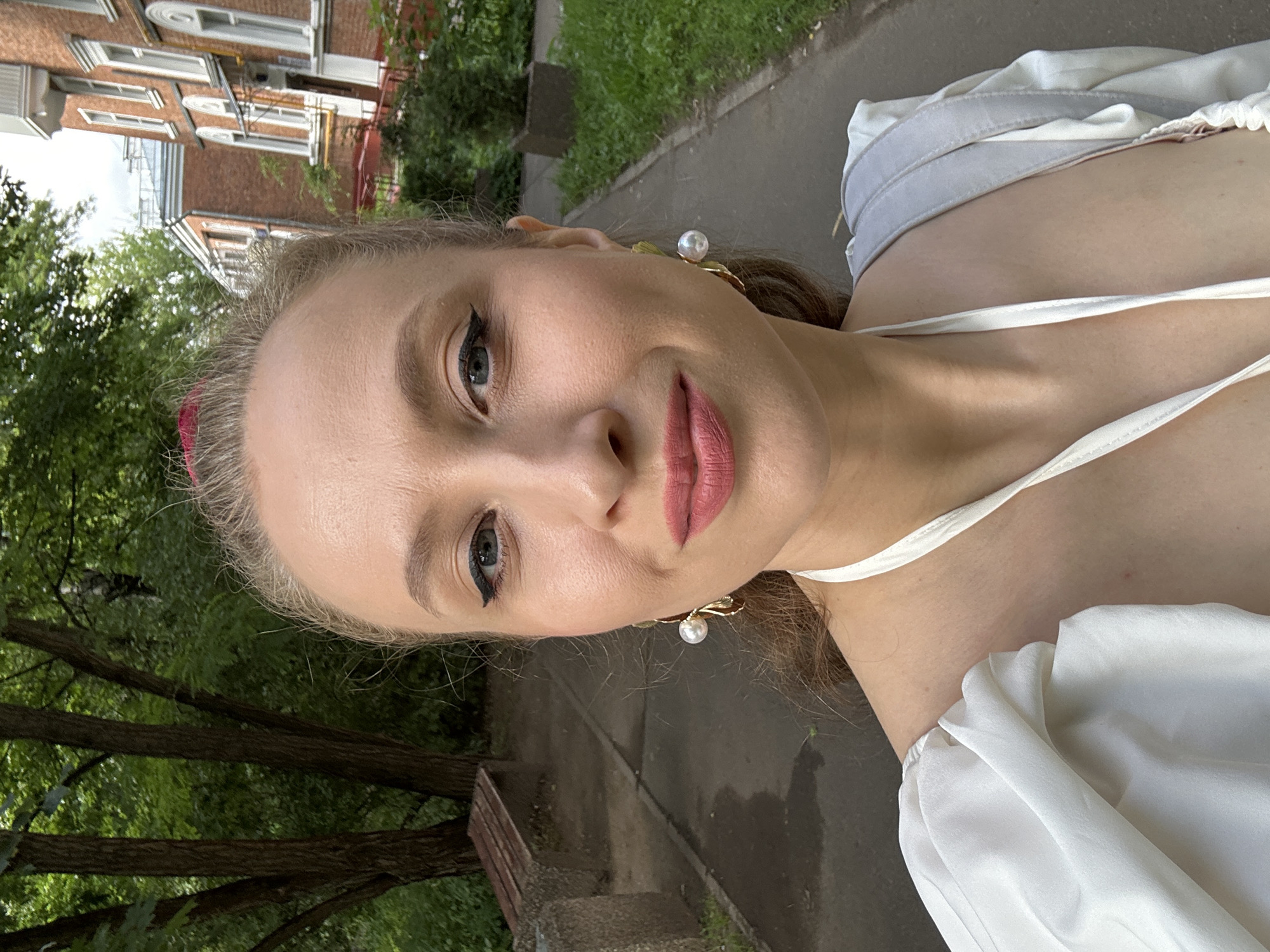 This media file is a vertical, close-up selfie of a young woman taken outdoors. She is facing the camera with her head tilted to her right. The woman has blonde hair and is wearing makeup, including eyeliner and lipstick. She is also wearing a white top with delicate straps and a pearl earring on her right ear. The background shows a brick building with windows and trees with green foliage, indicating an urban or suburban setting with vegetation. There are hints of a paved walkway and grass. The lighting suggests it is daytime, and the weather appears to be pleasant, with the sun illuminating the scene. The overall impression is casual and summery. No text is visible in the image. Based on the context provided, the location is Moscow, Russia.