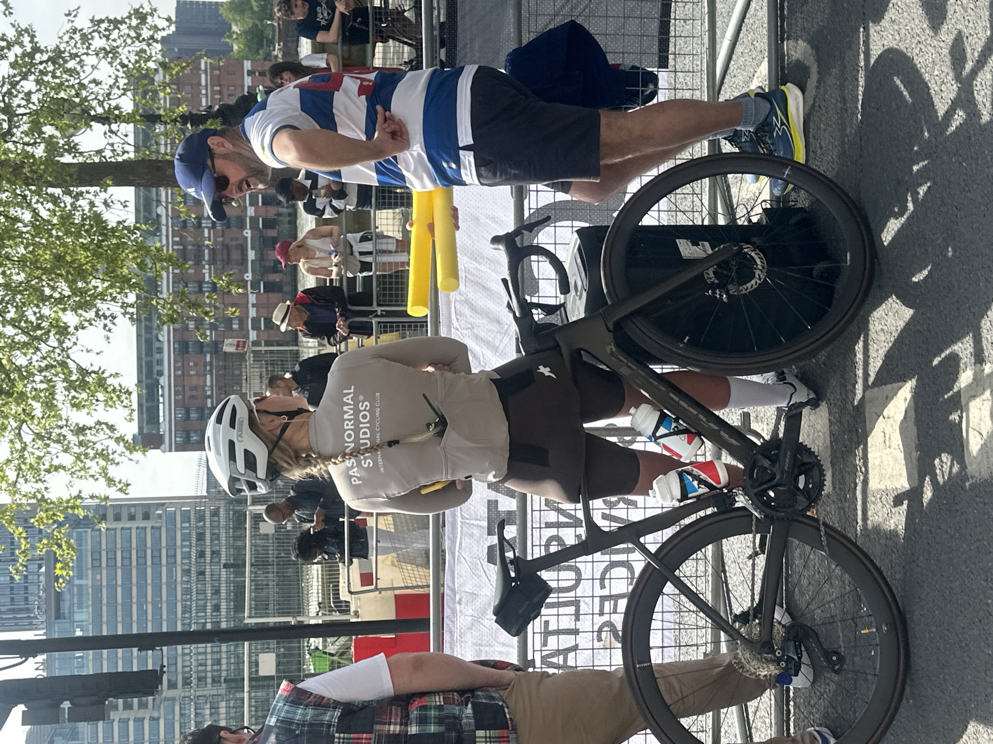 The image shows a scene in London, United Kingdom. A woman in cycling gear, including a white helmet and a "Pas Normal Studios" jersey, is standing next to a black road bicycle. The bike has water bottles mounted on its frame. Several other individuals are present: a man in a blue and white striped shirt, shorts, and a blue cap is standing nearby, and others are visible in the background, some behind a metal fence. The background includes city buildings and trees. Two yellow foam pool noodles are held by one of the figures. The setting appears to be outdoors on a sunny day.