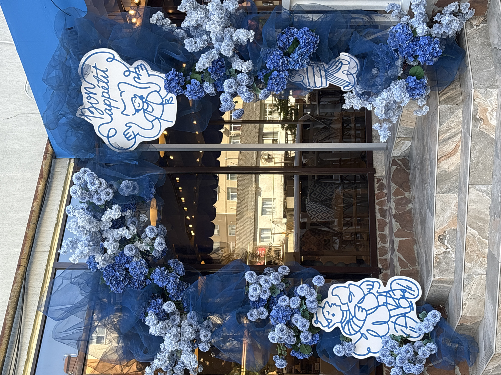 A decorative display of blue and white flowers, interspersed with sheer blue fabric, frames the entrance of a building in Sochi, Russia. The floral arrangement, featuring various shades of blue from light to dark, wraps around what appear to be glass windows, creating a vibrant archway.
Two prominent white, cloud-shaped signs with blue outlines are integrated into the floral decor. The sign on the left features the stylized text "Bon appétit" alongside a cartoon-like figure, possibly an angel or cherub, with wings and a smiling face, rendered in blue line art. The right sign also displays blue line-art figures in a joyful, interlocked pose, suggesting a celebratory or playful theme.
The building's lower facade is constructed with textured, earthy-toned stone or tile. Reflections in the large glass windows reveal glimpses of other urban buildings with multiple windows, indicating a city street setting. The scene is brightly lit, suggesting a clear day. The overall aesthetic is inviting and festive, likely adorning a cafe or restaurant entrance.