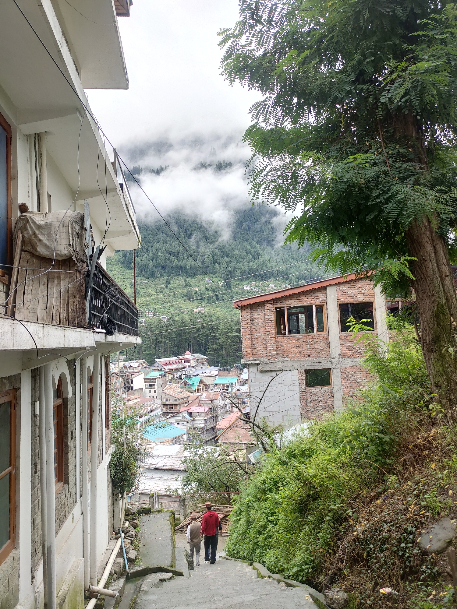The image shows a view of Siyal, India, from a hillside. In the foreground, on the left, is a white building with a balcony. To the right of the building, two men in traditional attire walk down a stone staircase. Beyond the staircase, a cluster of buildings with colorful roofs forms the town. The town is nestled in a valley, with a dense forest covering the surrounding mountains. The sky is overcast, with clouds obscuring the peaks.