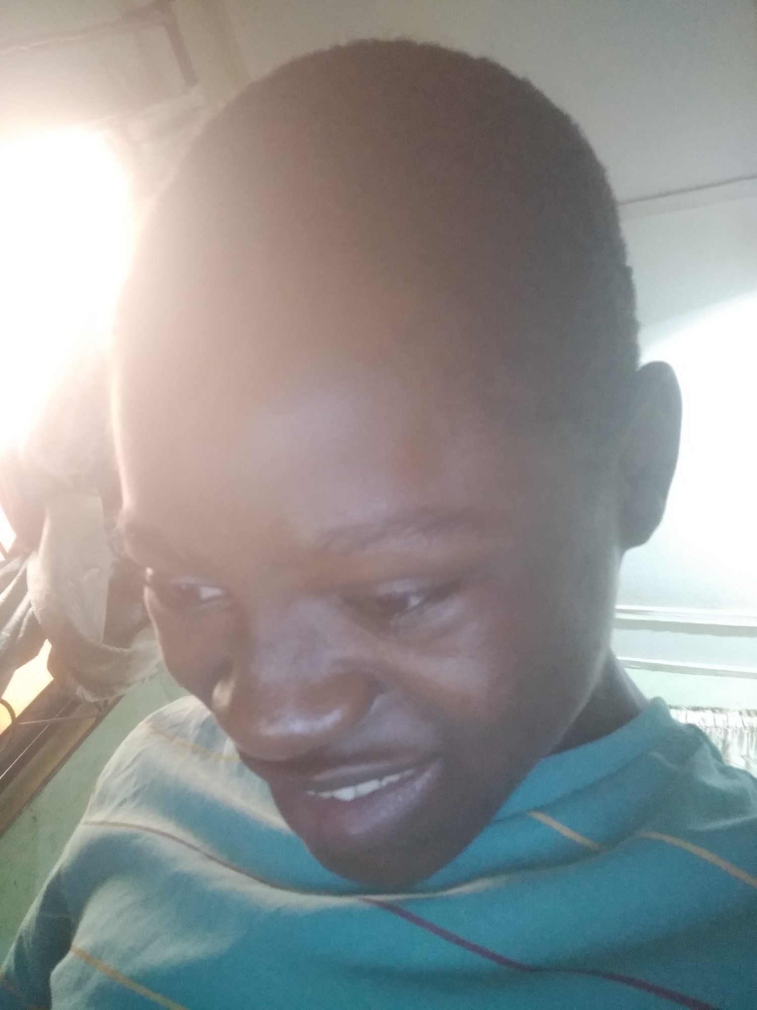 A person with dark skin and short hair is looking downwards, with a slight smile. They are wearing a teal striped shirt. The background is blurry, but appears to be an interior space with light coming from a window or artificial light source. The location and city are unknown.