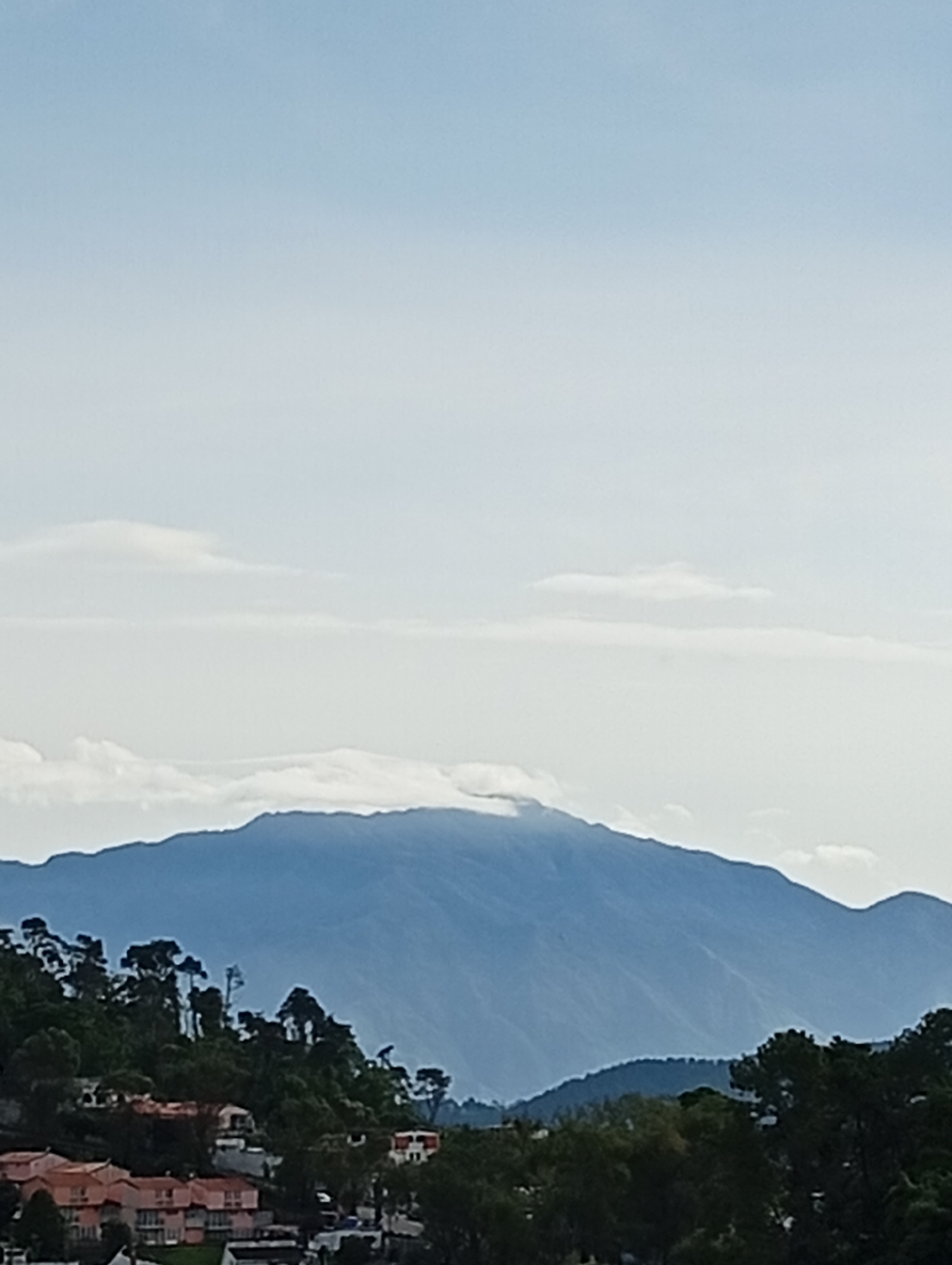 The image shows a hazy mountain landscape viewed from a residential area in San Antonio de Los Altos, Venezuela. The sky is a pale blue with some light clouds. In the foreground, there are buildings with reddish-orange roofs and several trees. The middle ground features a densely wooded hillside. The background is dominated by a large, blue mountain range, with clouds obscuring its peak. The overall atmosphere is serene, with no people or animals visible. The scene appears to be a scenic view of the natural environment surrounding the city.