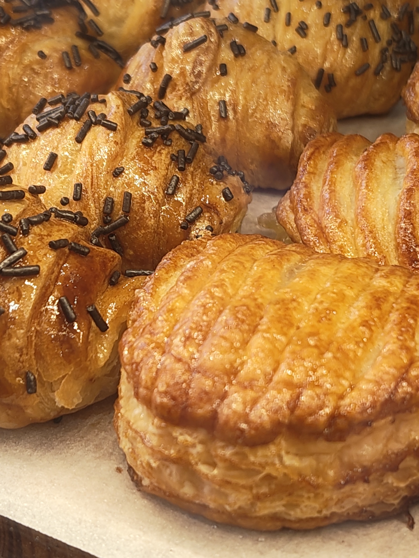 The image shows a close-up of several pastries, likely on display in a bakery. The pastries are golden brown and appear to be made of puff pastry. Some are topped with chocolate sprinkles. The pastries are resting on a white surface that is possibly parchment paper. The specific location within Dénia, Spain, is not discernible from the image alone, but they may be for sale at a bakery, cafe, or pastry shop. No people are visible. The focus of the scene is the assortment of pastries and their appealing presentation.