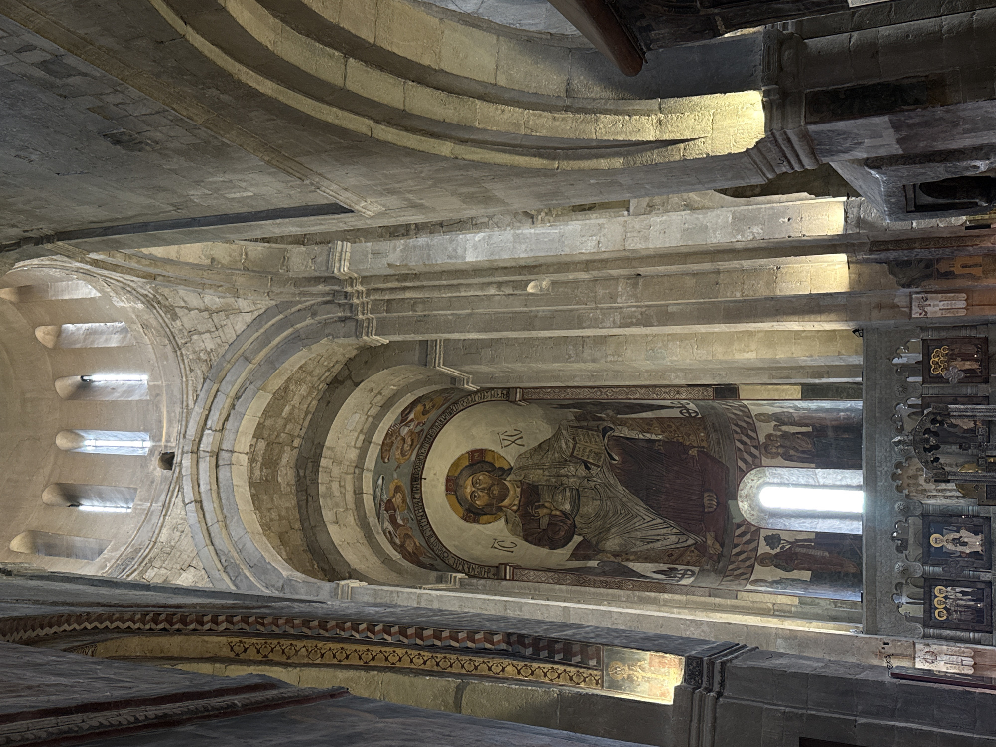 This image captures the interior of the Svetitskhoveli Cathedral, located in Mtskheta, Georgia. The camera is pointed upwards, offering a view of the arched ceiling and the walls above. The architecture is constructed from light-colored stone. Frescoes, including one prominently featuring a religious figure inside a circular frame, adorn the upper walls. The space is illuminated by natural light coming through window openings. The environment consists of stone construction, artistic frescoes, and architectural arches.