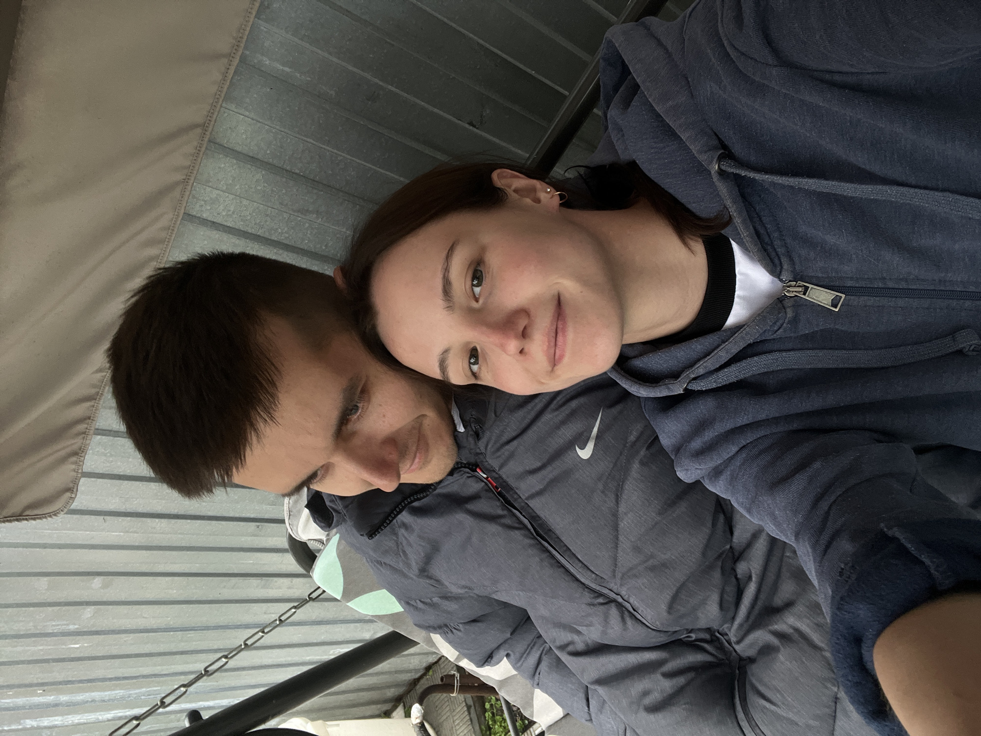 The image portrays two young adults, a man and a woman, seated closely together on an outdoor swing. The woman, positioned in the foreground and slightly to the right, has dark brown hair, fair skin, and is looking directly at the camera with a subtle smile. She wears a dark blue hooded zipper sweatshirt over a white t-shirt with a black collar band, and has a small gold hoop earring and a stud in her left ear. The man, seated behind and to the left of the woman, has short dark brown hair, fair skin, and light-colored eyes, and is smiling gently while looking towards the woman's right. He is dressed in a dark grey puffer jacket featuring a white Nike swoosh logo on the left chest, with a light-colored, patterned shirt visible underneath.
They are situated on a swing set, indicated by a black metal support chain on the left, a segment of a mint green and grey patterned cushion beneath the man, and a beige fabric canopy visible overhead. The background consists predominantly of a light grey corrugated metal fence or wall with vertical ridges. In the lower-left corner, a small patch of green vegetation and some light-colored paving stones are partially visible. The scene is illuminated by soft, diffused light, characteristic of an overcast day. While the location is specified as the city of Novocheremshansk, Russia, the visual content of the image does not contain specific identifying landmarks or features to confirm this geographical detail.
