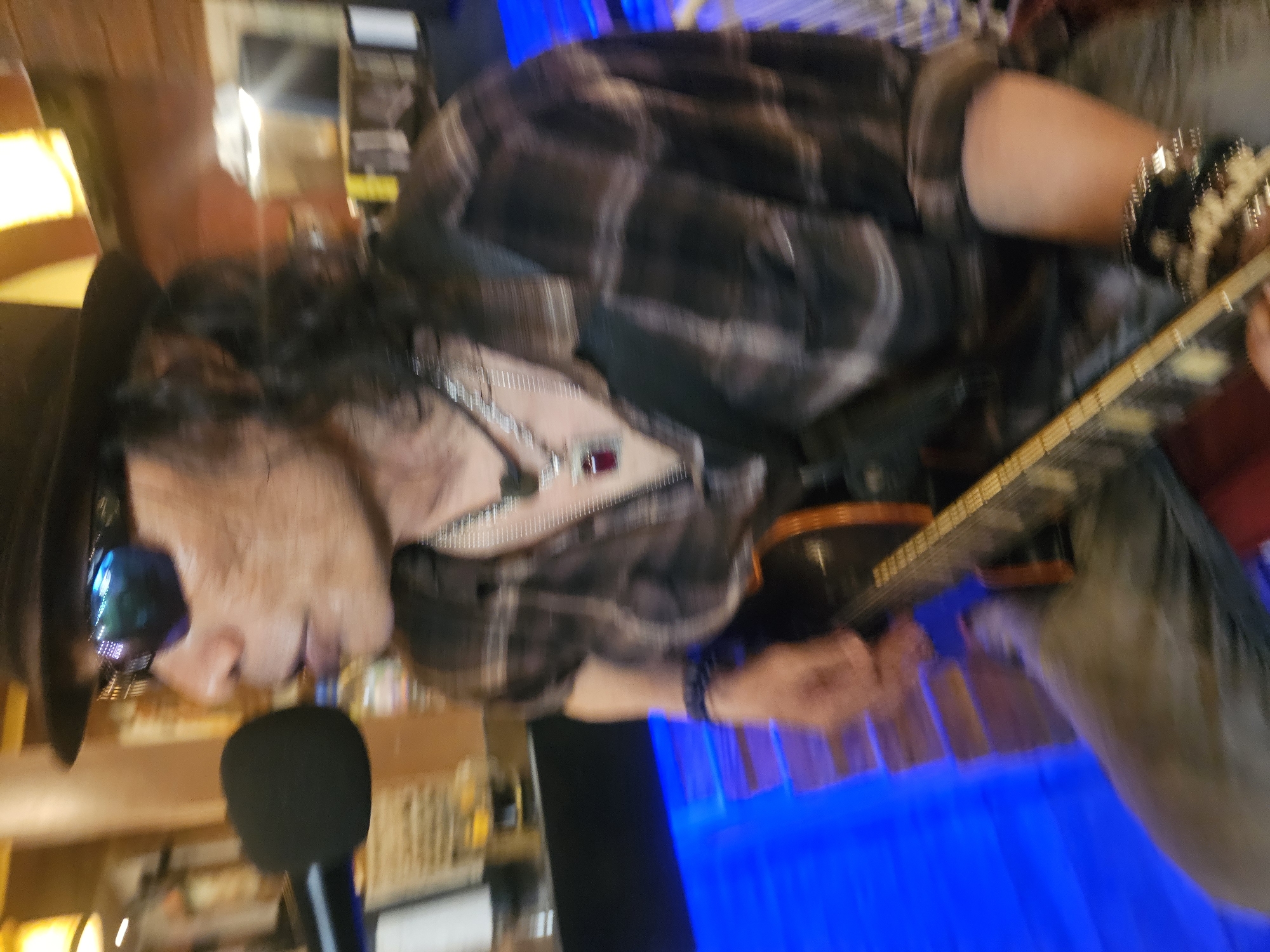A medium shot captures a person wearing a fedora, sunglasses, and a plaid shirt, playing a guitar. The person is positioned towards the right side of the frame, with their head and upper body visible. A microphone is located to the left of their face. The background is blurred, but appears to be an indoor setting with wooden elements and blue lighting. The overall impression is that of a musician performing in a venue. The location is specified as Denpasar, Indonesia.