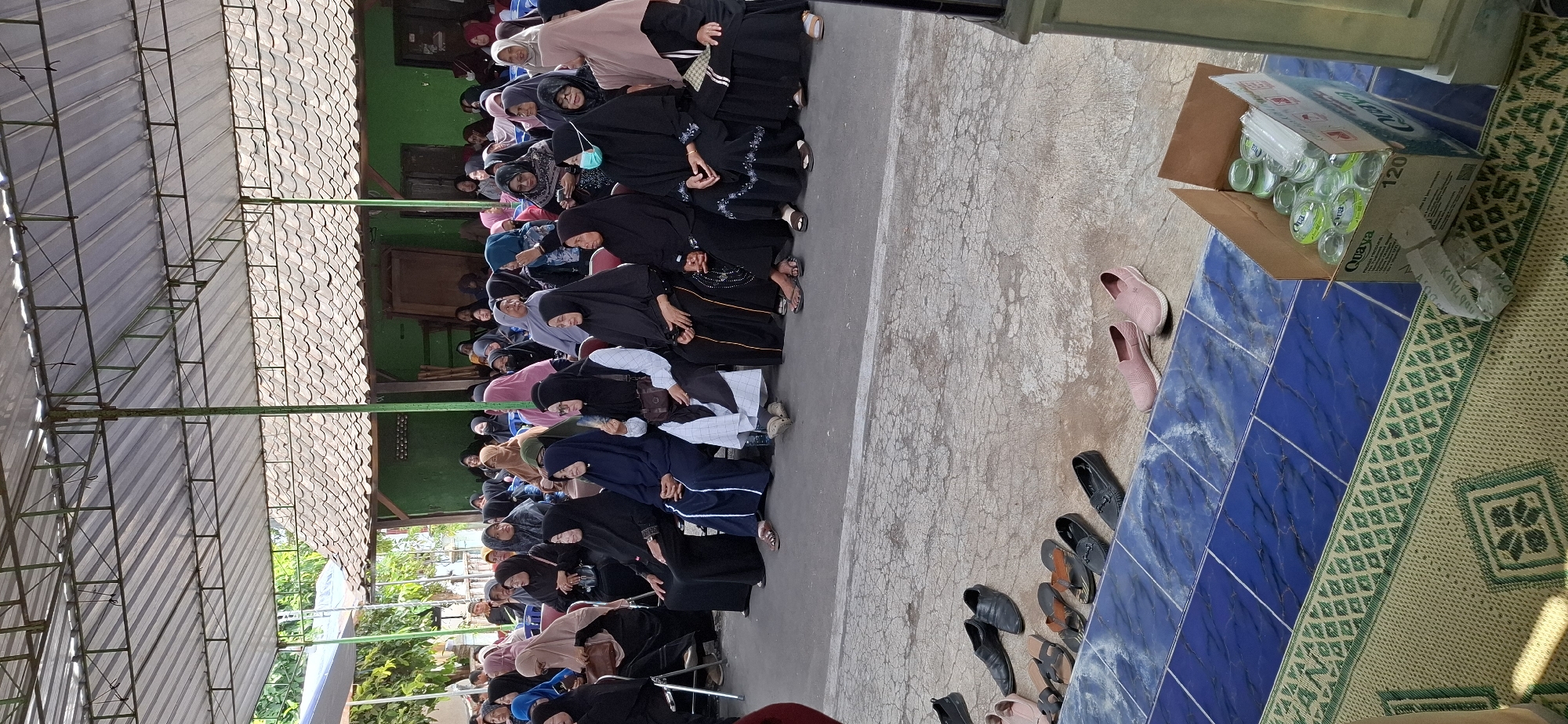 In Gentan, Indonesia, a group of women, most wearing hijabs and traditional attire, are seated in rows under a covered outdoor area. The structure has a corrugated metal roof supported by a metal frame. Behind the seated individuals, a building with a tiled roof and green walls is visible. The ground in front of the seating area is a concrete surface with some tiled sections and a woven mat. Several pairs of shoes and sandals are scattered on the concrete. A cardboard box filled with bottled drinks and straws is placed on the mat. The scene appears to be an outdoor gathering or event.