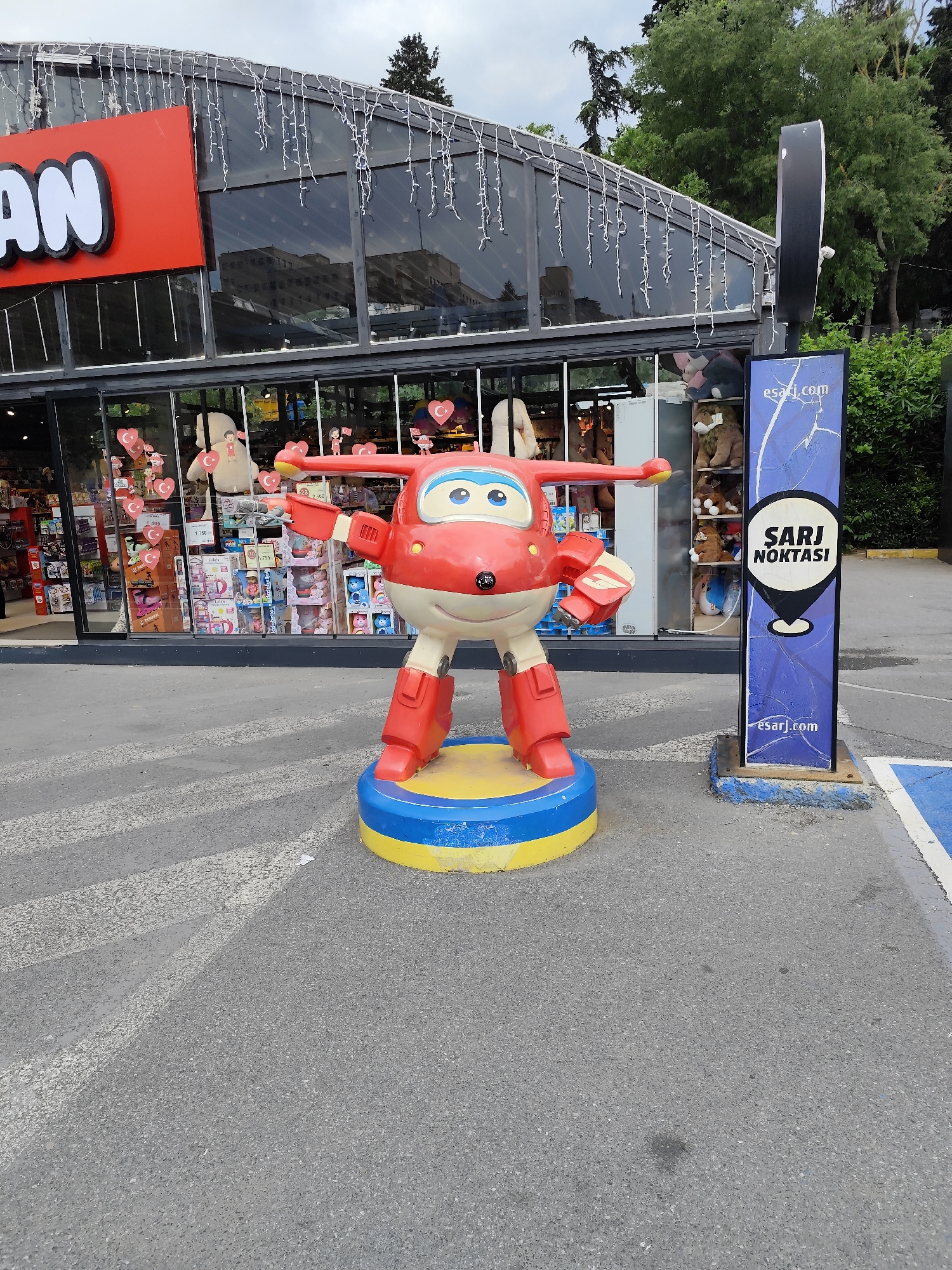 The image depicts a figure of Jett from the children's show "Super Wings" standing on a platform in front of a store in Sarıyer, Turkey. The figure is red, white, and yellow. It stands on a blue and yellow platform. A sign reads "Sarı Noktası." The storefront is visible in the background, with toys displayed in the windows. Overhead, string lights adorn the building. The ground appears to be asphalt. The scene is set outdoors during daylight hours.