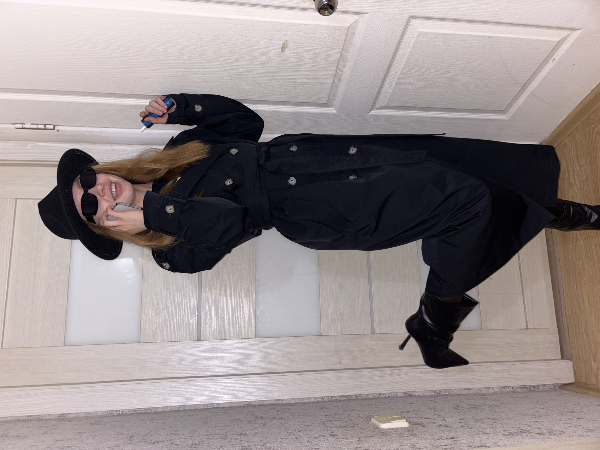 The image depicts a young woman posing indoors, shown rotated 90 degrees clockwise as if lying horizontally against a white wooden door in Perm, Russia. She has long, straight blonde hair and is smiling broadly, revealing her teeth. Her attire includes a black wide-brimmed hat, black sunglasses, a long black double-breasted trench coat with a belted waist and light-colored buttons, black pants, and black high-heeled ankle boots.
She is engaged in a casual pose, holding a light-colored smartphone to her ear with her left hand and a blue and white electronic vaping device in her right hand. The background features a white paneled door with a silver door handle visible at the top. The floor transitions from light wood laminate on the right to a gray carpet or rug on the left, separated by a light wooden baseboard. A small, light-colored rectangular object, possibly a button or a piece of plastic, lies on the carpet near her right foot. No discernible text is visible in the image. The scene is brightly lit, typical of an indoor environment.