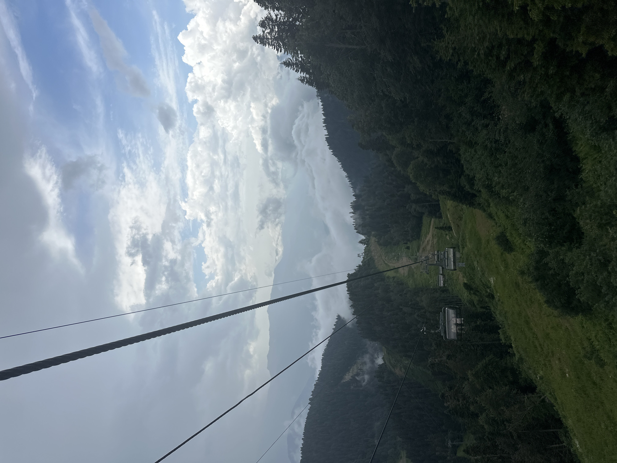 The image shows a cable car ascending a steep, verdant mountainside in Estosadok, Russia. The cable car cabins, suspended from thick cables, carry unseen passengers up the slope. Dense evergreen forests cover the mountains, with patches of grassland visible along the cable car path. In the distance, rolling mountains are partially obscured by a layer of fluffy white clouds, creating a sense of depth and elevation. The sky above is a mix of bright blue and scattered grey clouds, suggesting a dynamic weather system. The overall impression is one of natural beauty and outdoor recreation in a mountainous region.
