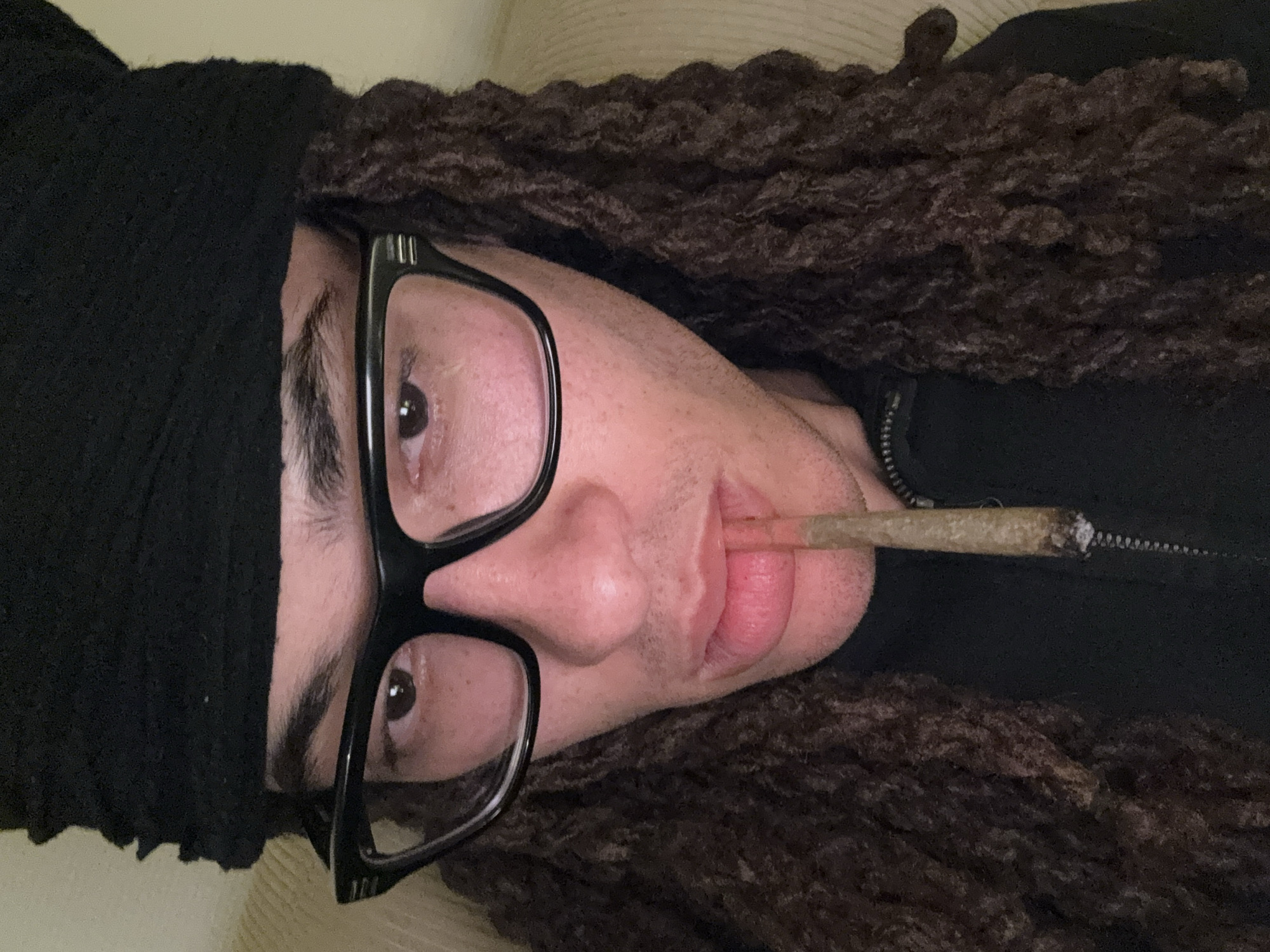 This close-up portrait, oriented sideways, features a person with a light to medium skin tone, framed against a plain, light-colored background that appears to be an indoor setting. The individual is wearing a black beanie and black-rimmed glasses, which obscure part of their eyes. Dark brown, textured dreadlocks or braids are visible, extending from under the beanie. The person is holding a lit joint between their lips, with visible ash at the tip, suggesting they are in the process of smoking. A portion of a dark jacket with a zipper can be seen on their right side. Their gaze is directed towards the camera, and their expression is neutral. The lighting is soft, highlighting facial features like the nose and lips. No specific location cues are visible within the image itself to confirm the provided context of Paris, France. There is no visible text.