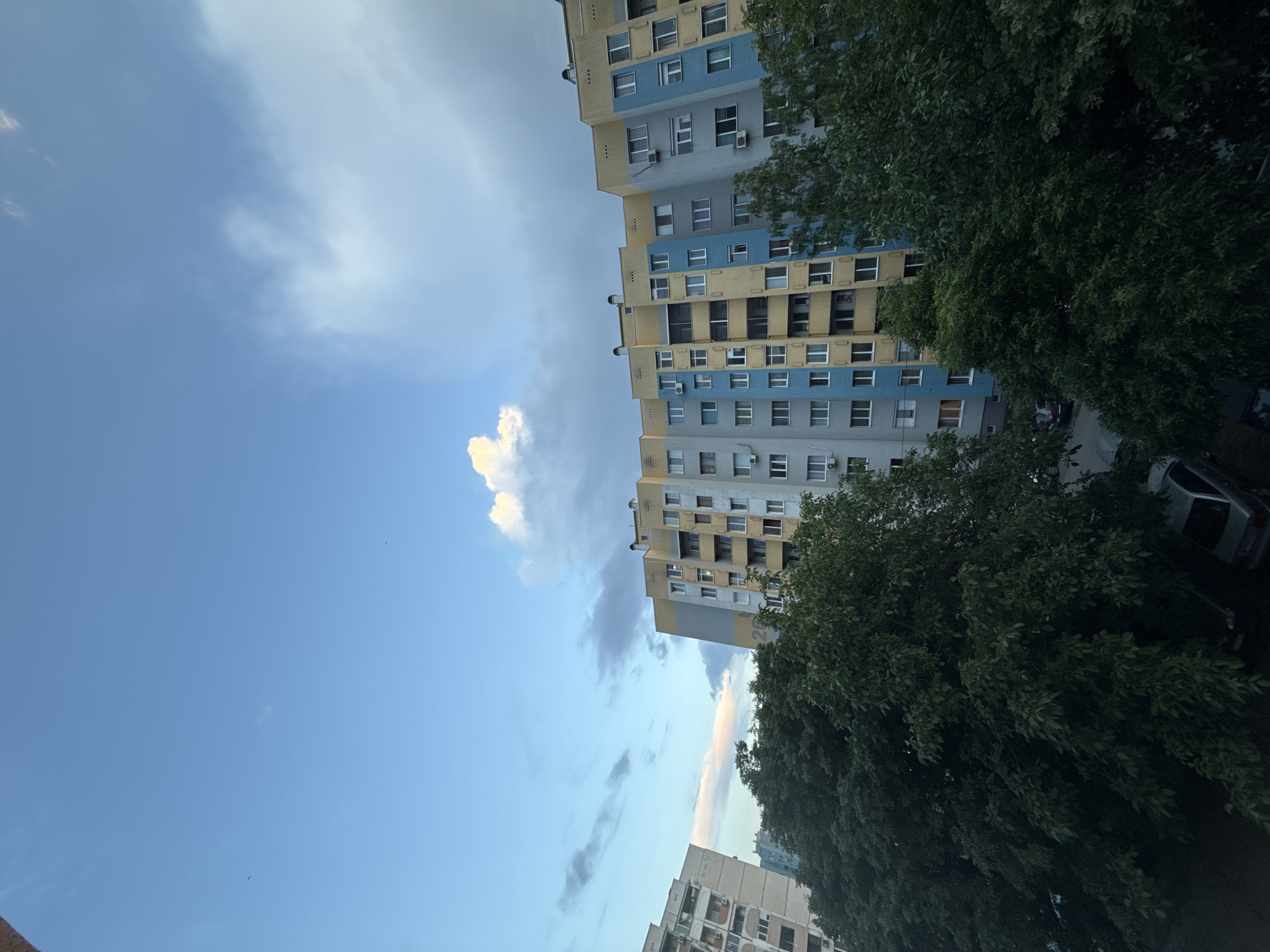 The image depicts a view of a multi-story building in what appears to be a residential area. The building is painted in blue, gray, and yellow. The building's architecture is characterized by many windows. Trees are partially obscuring the building. The sky is partly cloudy with blue patches. Based on the location and the color scheme of the building, it is in Stara Zagora, Bulgaria. A car parked at the base of the building.