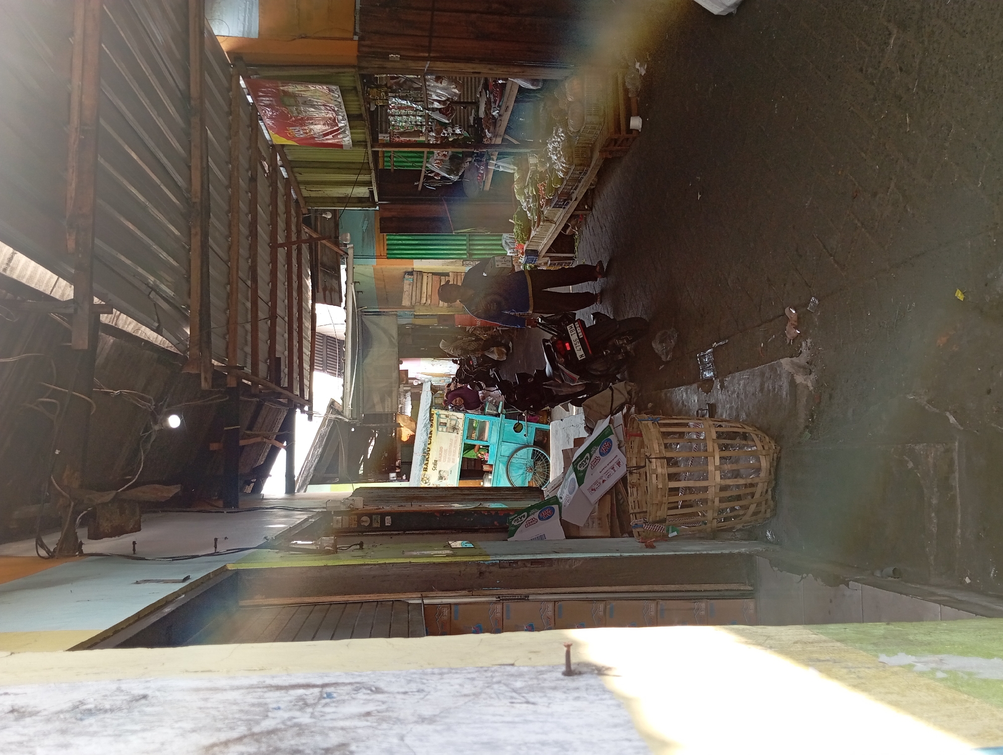 The image depicts a street market in Kepulungan, Indonesia, with stalls lining a narrow, covered walkway. The market appears to be an open-air establishment, with corrugated metal roofs supported by wooden beams. The ground is paved with dark, uneven stones, and there are some puddles of water present, suggesting it may have recently rained or there is poor drainage.
On the right side of the walkway, several market stalls are visible, displaying a variety of goods. One stall is laden with fresh produce, including what appear to be onions, garlic, and various vegetables arranged in crates. Another stall further back has meat or poultry hanging from hooks. A person, wearing a blue long-sleeved shirt and dark pants, is standing near the produce stall, holding a white box and looking towards the left.
On the left side of the walkway, a blue cart with a bicycle wheel is parked. In front of it, there are some discarded cardboard boxes and a large woven basket. Further down the walkway, a motorcycle is parked. Another person, wearing a headscarf and a dark dress, is partially visible behind the blue cart.
The overall atmosphere is one of a bustling, yet somewhat weathered, local market. The lighting is natural, with sunlight streaming in from the top, creating bright patches and shadows. The buildings on either side of the walkway are simple, with painted walls, and appear to be part of the market's infrastructure. The environment appears to be somewhat cluttered with goods, packaging, and debris.