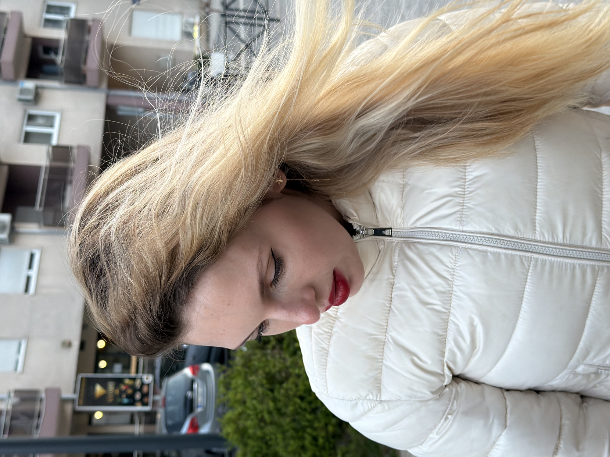 An outdoor, slightly tilted portrait shot features a young woman with long, wind-swept blonde hair, appearing to look downwards or with her eyes gently closed. She is positioned on the right side of the frame, facing slightly left. Her hair is a vibrant blonde with subtle darker undertones at the roots, and numerous fine strands are visible, suggesting movement. Her complexion is fair, accented by dark eyeliner and striking red lipstick. A small, golden, star-shaped earring adorns her left ear. She is dressed in a cream or off-white quilted puffer jacket with a prominent silver zipper.
The background portrays an urban environment consistent with Budva, Montenegro. Multi-story buildings with windows, balconies, and external air conditioning units are visible under what appears to be soft daylight. Below the buildings, a street scene includes a silver car partially visible on the left and a patch of green foliage. A commercial sign or billboard with lights and indistinct text can also be seen in the background, adding to the urban context. The overall impression is one of calm or contemplation.