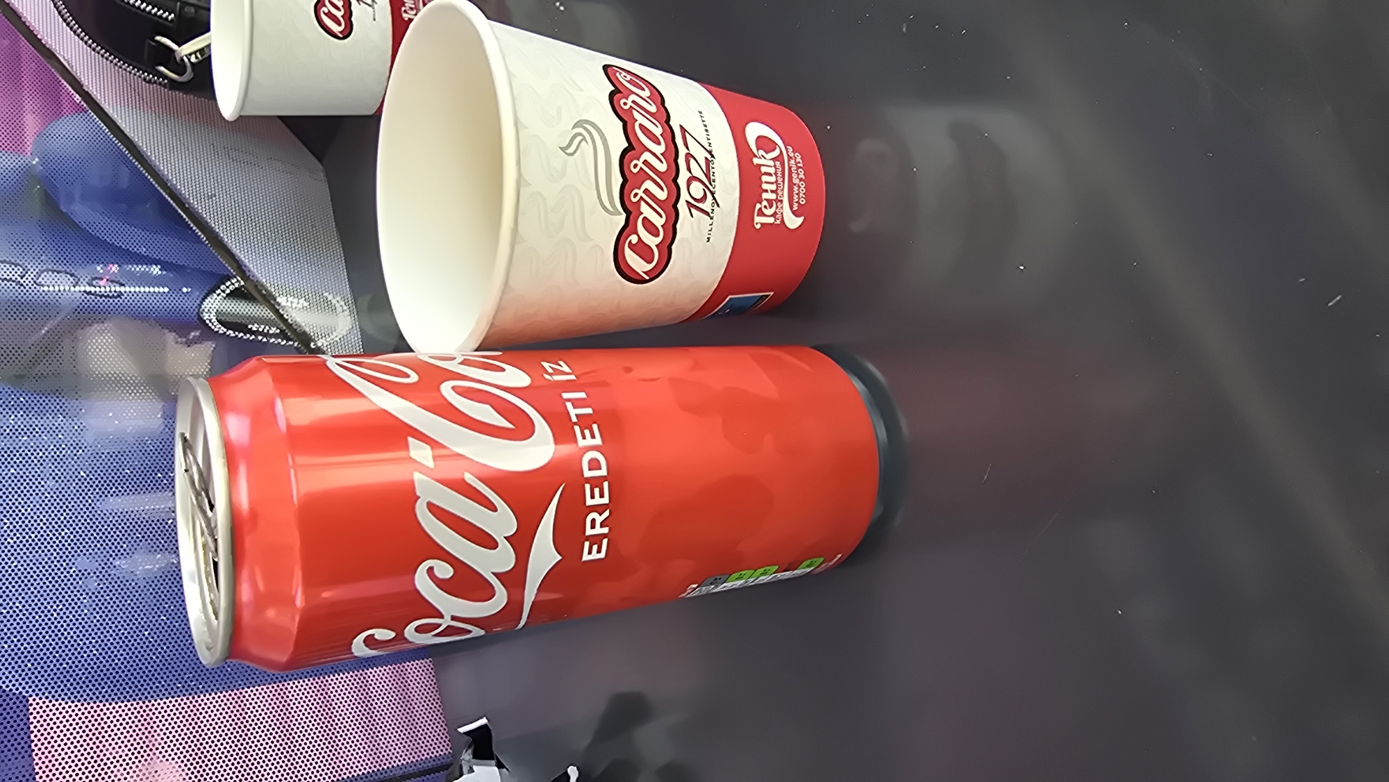The image depicts a close-up, eye-level view of a Coca-Cola can and a disposable coffee cup placed on a dark, reflective surface, likely a table. The Coca-Cola can is red with the iconic white logo and the Hungarian text "EREDETI ÍZ" (Original Taste) visible. Next to it, a white paper coffee cup features a red and white logo that reads "Carraro 1927" with smaller text below. The bottom part of the cup has a red band with white text that includes "Genik.eu" and a phone number. In the background, out of focus, is a textured blue surface with a mesh pattern, and a black object resembling a bag or a handle. The scene suggests a casual setting, possibly an outdoor cafe or a break area. There are no people visible in the image. The image does not provide any information or visual cues that would allow for the identification of its location in the city of Vratsa, Bulgaria.