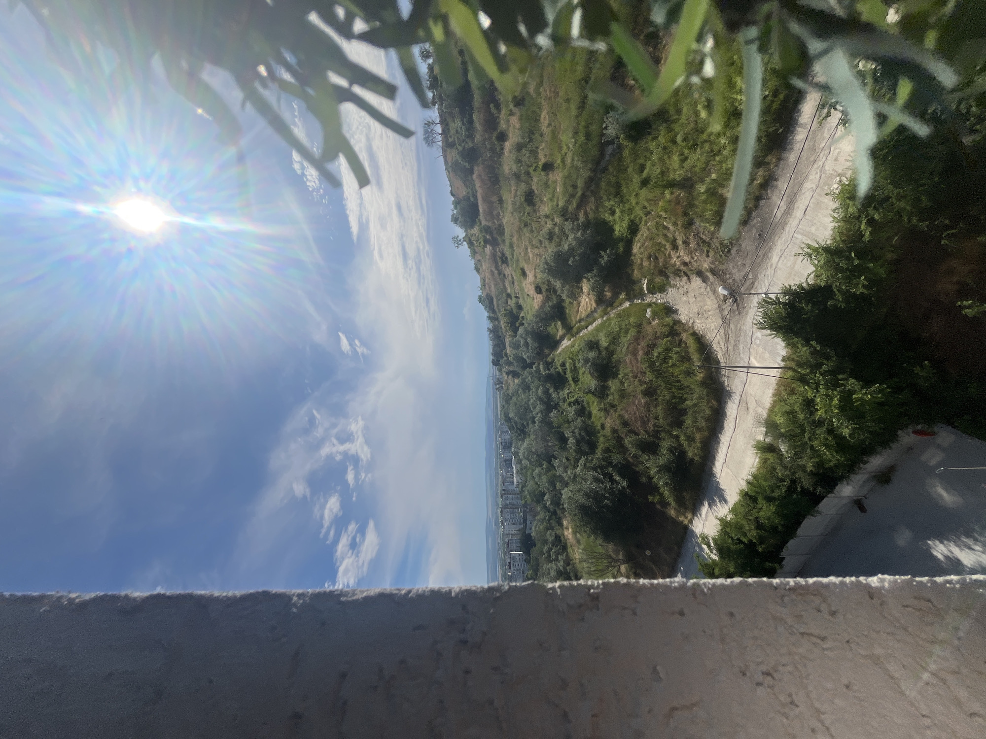 The image presents a sun-drenched vista from a high vantage point, looking out over a hilly landscape towards a coastal city, presumably Varna, Bulgaria. In the foreground, a rough, textured wall, likely part of a building, occupies the left side of the frame, casting a shadow that partially obscures the view. Beyond this, a sunlit, partly cloudy sky dominates the upper portion of the image, with the sun prominently positioned, creating a lens flare effect.
The mid-ground reveals a verdant, undulating terrain covered in green shrubs and dry grass, with a dirt path winding its way through the landscape. A few scattered trees punctuate the hill. In the distance, a cluster of multi-story buildings, characteristic of a residential area, are visible near the horizon, suggesting the presence of the city. The overall impression is one of a bright, clear day, with nature encroaching on a developed area. The foreground wall and the hint of vegetation in the upper right corner suggest an outdoor setting, possibly a balcony or rooftop overlooking the scene. The presence of the buildings and the general topography are consistent with a coastal city like Varna, which is known for its hilly landscape leading down to the Black Sea.