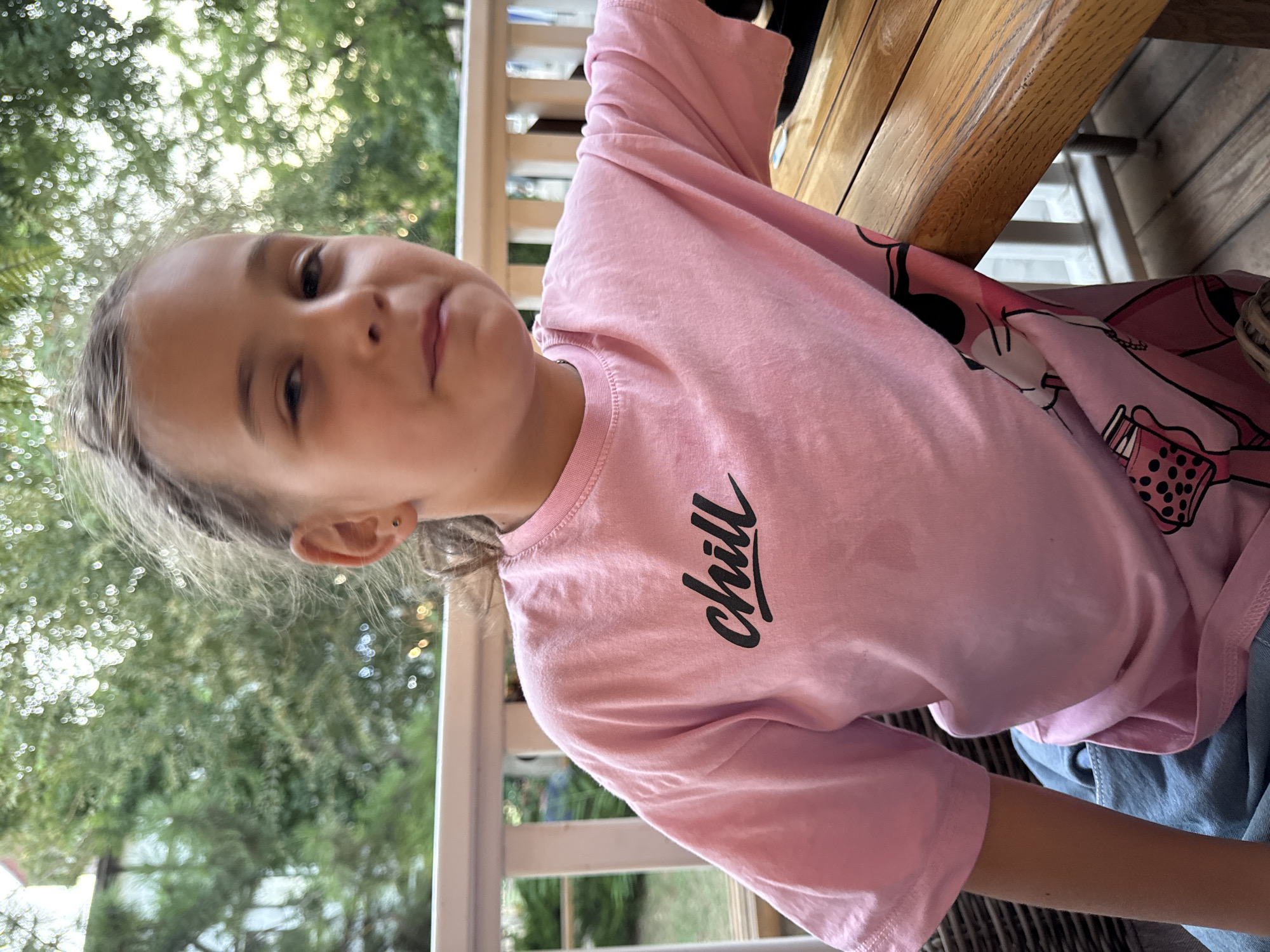 A young girl is shown from the chest up, looking towards the camera with a slightly open mouth. She has light brown hair pulled back, and is wearing a pink t-shirt with the word "chill" in black cursive lettering across the chest. She also appears to be wearing denim shorts. The background is blurred but shows green trees and a white railing, suggesting an outdoor setting, possibly a porch or balcony. A wooden table is to the right of the girl, partially obscuring her from view. The lighting suggests it is daytime, possibly late afternoon. The girl's expression could be interpreted as playful or surprised.