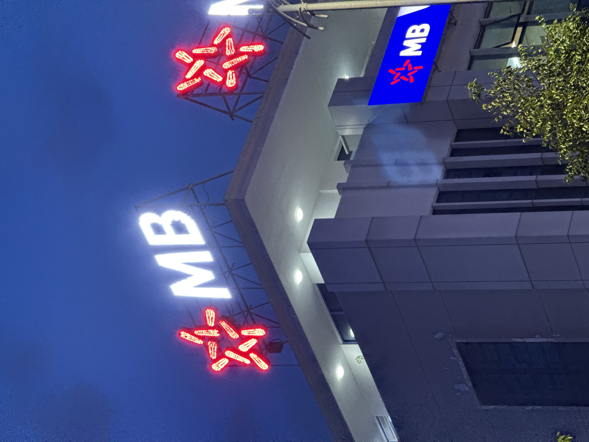 The image shows the exterior of an MB Bank building in Nha Trang, Vietnam, at night. The building is modern, with a grey facade and large windows. A prominent sign with the MB logo and the letters "MB" in white neon lights is visible on the rooftop. Red neon lights in the shape of a star also adorn the rooftop. Another blue sign with the MB logo and "MB" text is mounted on the side of the building. A tree with green foliage is partially obscuring the lower portion of the building. The sky is dark blue, indicating it is nighttime. No people are visible in the image.