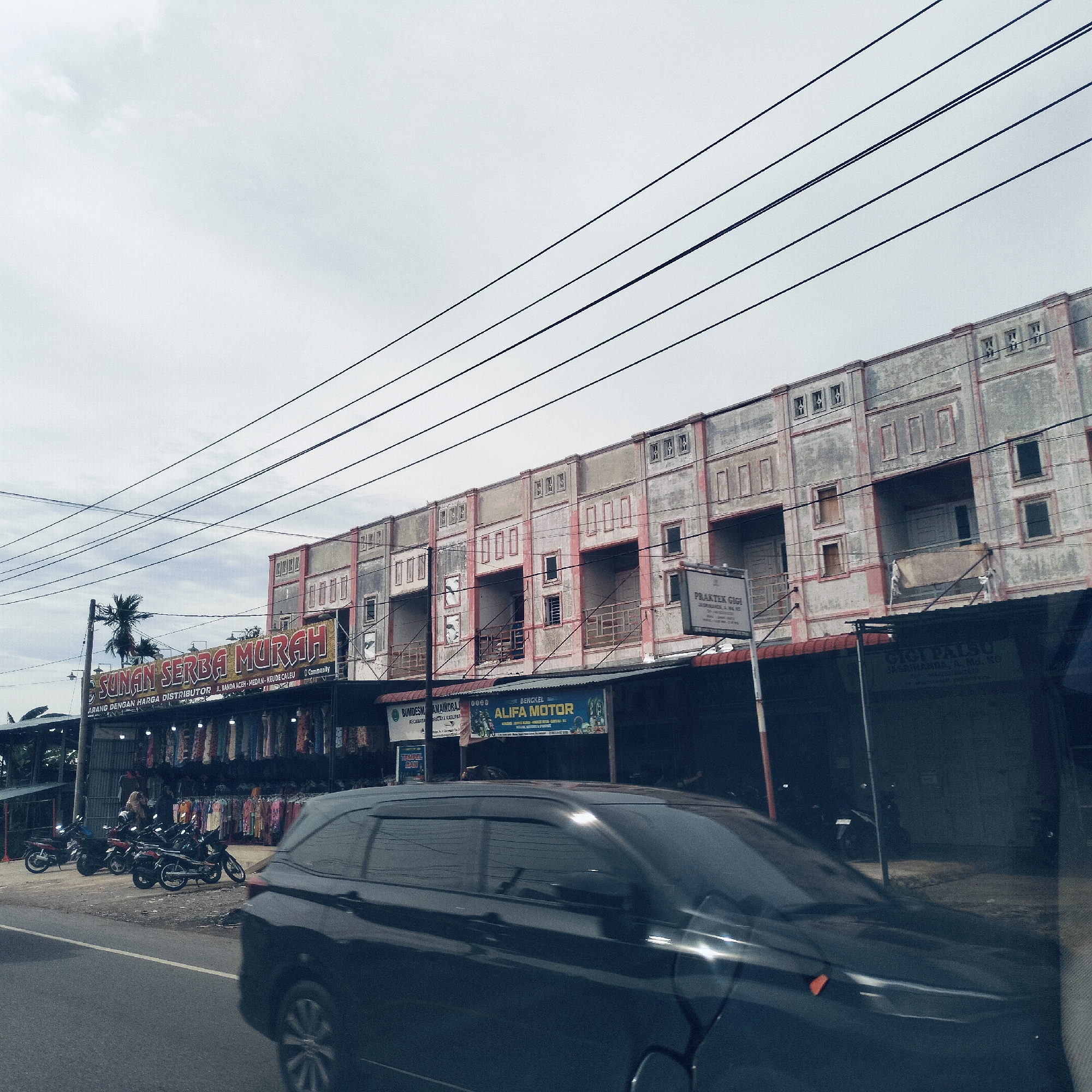 This image captures a daytime street scene in Teungoh Baro, Indonesia, under an overcast sky. The prominent subjects include a row of two-story concrete buildings lining the right side of the road, featuring multiple balconies and visible signs of wear with peeling paint. Overhead, numerous power lines crisscross the frame against the pale sky.
In the foreground, a dark-colored car, possibly an SUV or MPV, is captured in motion, its body blurred, suggesting movement. On the left side of the road, several motorcycles are parked near a shop. This shop, identified by a large yellow sign, displays an array of clothing on racks outside. A person is partially visible near these clothes racks.
Visible text on various signs provides commercial and service information:
* "SUNAN SERBA MURAH" with additional text "ARANG DENGAN HARGA DISTRIBUTOR JL. BANDA ACEH MEDAN - KEUDE CALEU Community" for the clothing shop.
* "BENGKEL ALIFA MOTOR" for a motor repair shop.
* "PRAKTEK GIGI" and "GIGI PALSU JASRINANDA, A. Md. KG" for a dental practice.
* A partially visible sign reads "BUMDESM AMAINDRAJ KECAMATA PRATAYA KABUPAT".
* Another small sign says "TEMPEL BAN".
The scene depicts a typical commercial street with various local businesses, suggesting an active urban or semi-urban environment.