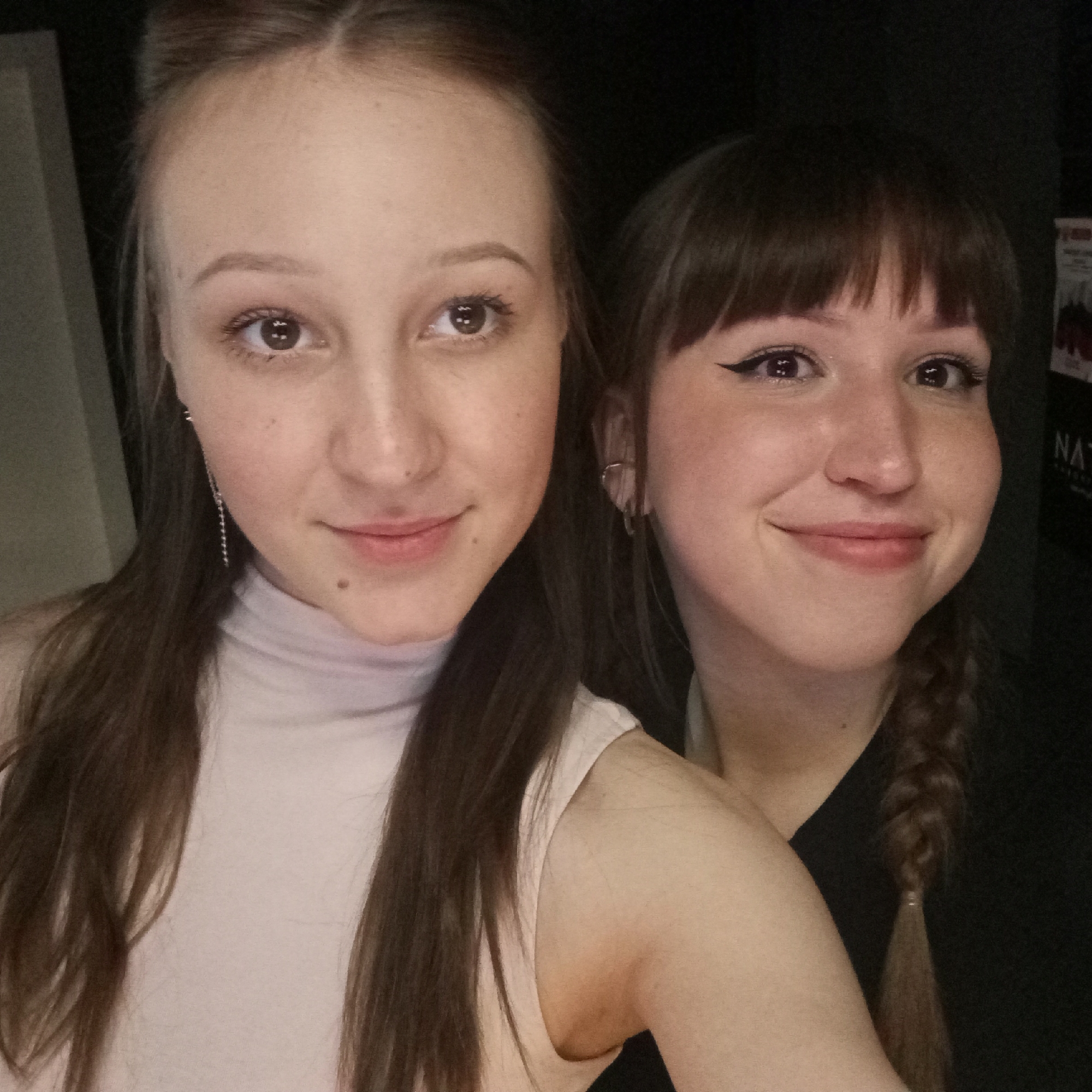 Two young women pose close together for a selfie. The woman on the left has long, straight brown hair, light skin, and is wearing a light pink turtleneck top. She has long, dangling earrings. The woman on the right has brown hair with bangs, eyeliner, and a braid hanging down her back. She is wearing a black top and small hoop earrings. The background is dark, possibly indoors, with what appears to be a poster partially visible in the top right corner, with the first two letters of the name visible as "NA". The location in the city of Chaikovsky, Russia is not discernible from the image.