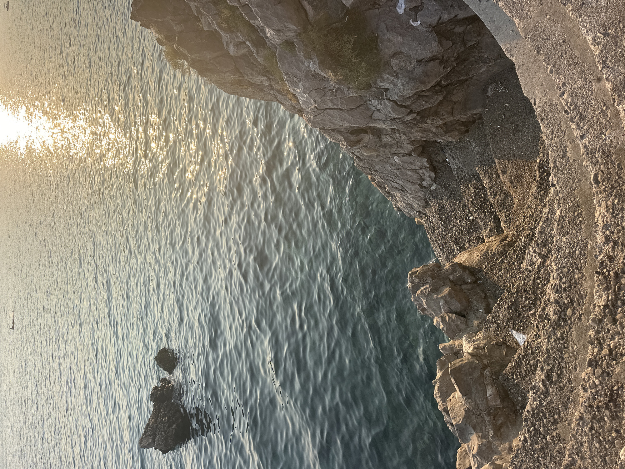 The image captures a rocky coastline with the Adriatic Sea in Budva, Montenegro. The water is a deep blue-green, with the sun glinting off its surface, creating a shimmering effect. Several dark, irregular-shaped rocks are visible emerging from the water on the left side of the frame. In the distance, a small boat with a person on it is visible.
In the foreground and right side of the image, a steep, rocky incline leads down towards the sea. There are man-made steps carved into the rock face, some of which are partially obscured by debris. The ground is covered in gravel and small stones. Vegetation, in the form of small green bushes, grows sparsely on the rocks. There are scattered pieces of white debris, possibly plastic waste or paper, visible on the rocks and steps. The overall scene suggests a natural, rugged environment with some human impact, particularly in the form of the steps and the debris. The lighting indicates it is likely late afternoon or early evening due to the warm, low-angle sunlight.