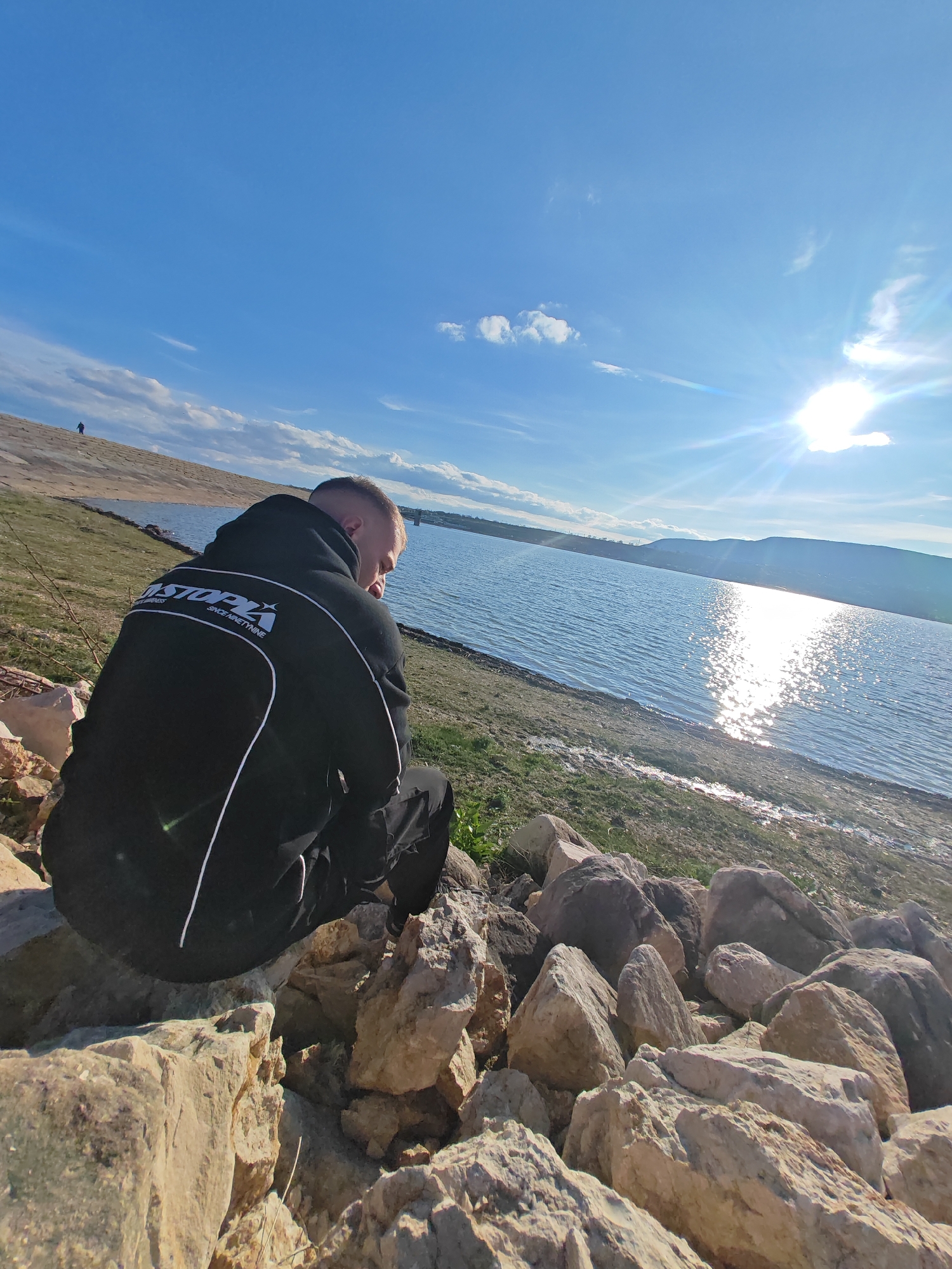 A person in a black hooded jacket with white stripes sits on rocks near a body of water. The person is positioned with their back to the camera, looking towards the water. The water reflects sunlight, creating a glare. Beyond the water is a shoreline with vegetation and a distant landmass. The sky is blue with scattered clouds. The location is believed to be near the city of Shumen, Bulgaria.