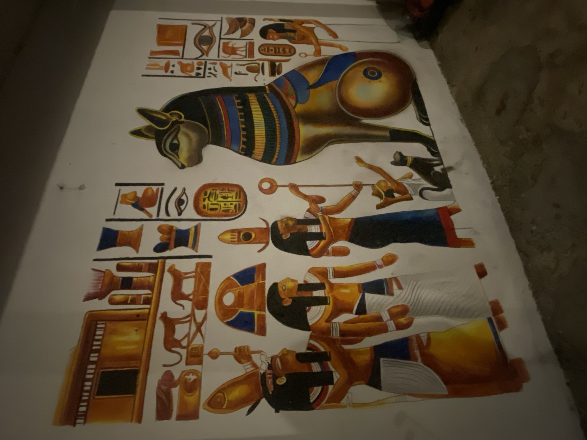 The image shows a depiction of ancient Egyptian art, likely a mural or a painted panel. The artwork displays various figures, hieroglyphs, and symbols typical of Egyptian art.
Subjects include:
* A stylized ram or bull with curled horns.
* Several human figures in profile, adorned with traditional Egyptian headdresses and attire.
* Hieroglyphic symbols organized in columns or rows.
Actions/Interactions:
* One human figure appears to be blowing a horn or trumpet.
Objects:
* The figures are holding objects like staffs or scepters.
* Egyptian-style furniture or architectural elements are represented.
Environment:
* The background appears to be a white or light-colored surface, suggesting a wall or panel.
Location:
The image is in the city of Baan Don Sai, Thailand.