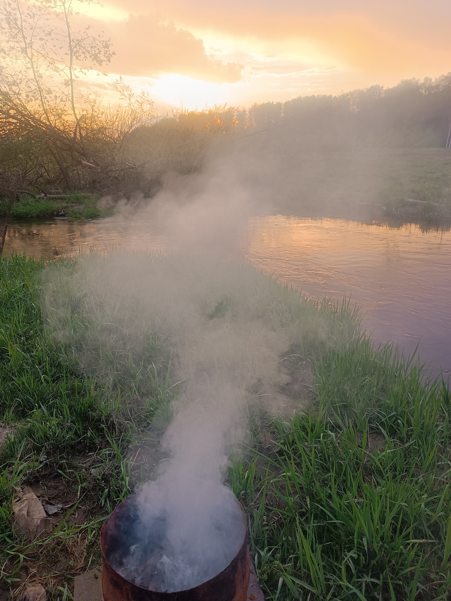 The image depicts a burn barrel with smoke rising, situated near a riverbank in Motyakovo, Russia. Green grass grows along the riverbank. The river reflects the orange-yellow hues of the sunset sky. Trees and foliage are visible in the background.