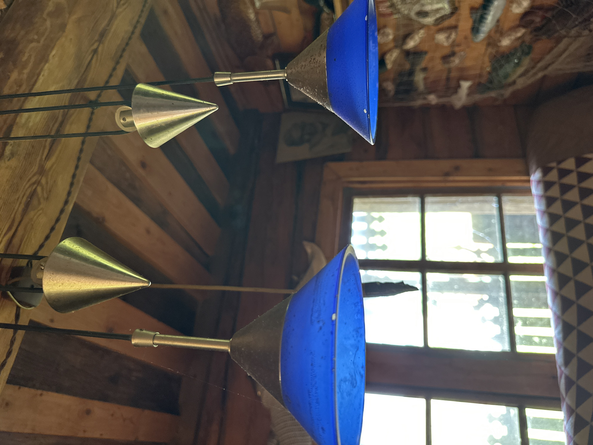 The image depicts an interior scene within a rustic log cabin, possibly in Norviliškės, Lithuania, characterized by exposed wooden beams and walls. Two pendant light fixtures, each with a metallic cone-shaped top and a vibrant blue glass shade, hang from the ceiling. One light fixture is closer to the foreground and is more prominently displayed, while the other is slightly further back and partially obscured. The blue shades appear to be speckled with dust or water droplets. Behind the lights, a window with multiple panes offers a view of the outdoors, with sunlight filtering through. A patterned upholstered piece of furniture, possibly a sofa or chair, is visible in the lower portion of the frame. The walls are decorated with various items, including a framed picture of a person and what appear to be decorative fish. The overall atmosphere is cozy and rustic.