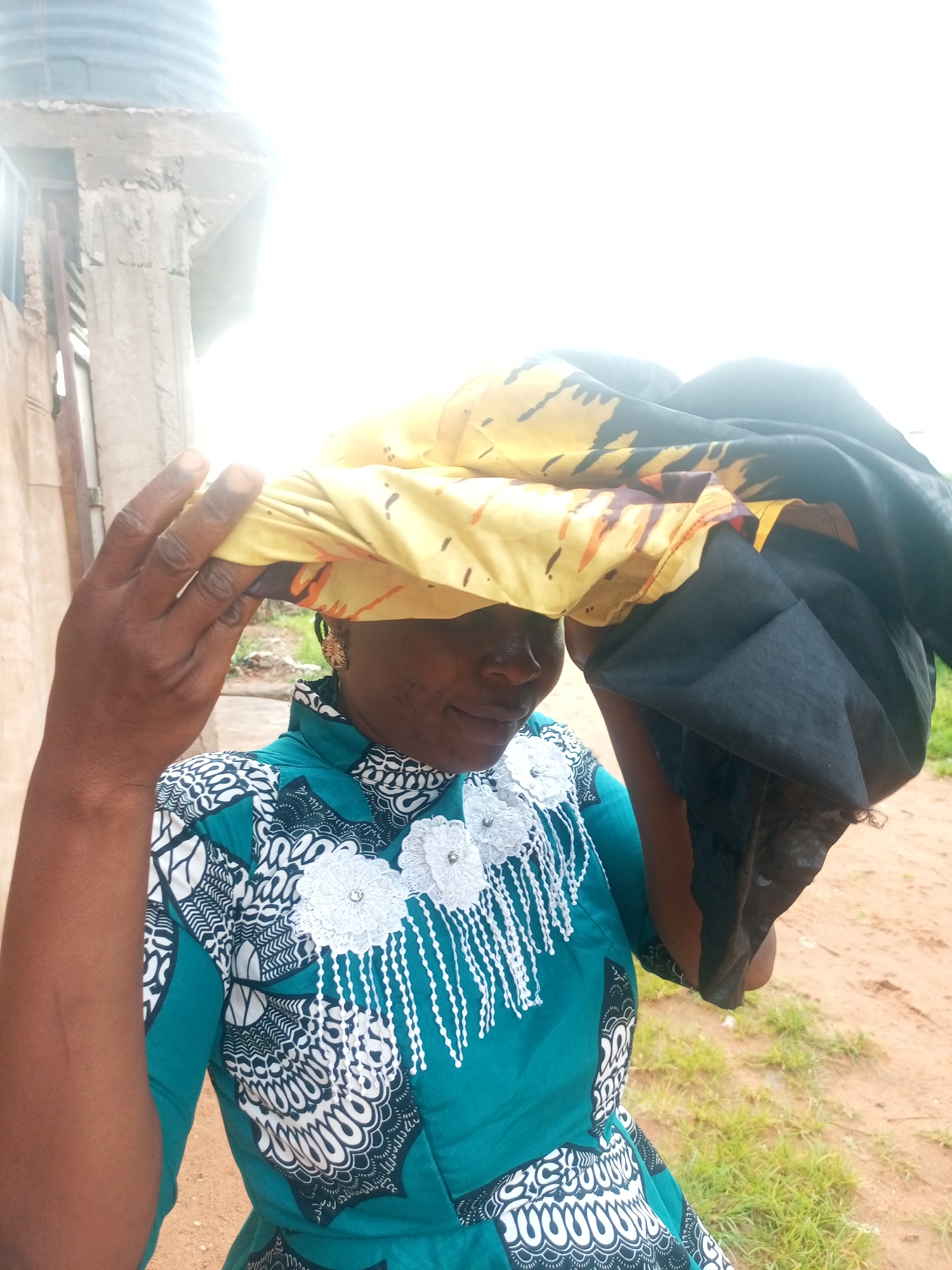 In an outdoor environment, a woman stands with a yellow and black patterned cloth draped over her head, obscuring her face. She is wearing a teal dress with a white floral and beaded embellishment around the neckline and down the front. The background shows a rough, light-colored wall and some green vegetation. A large, cylindrical object, possibly a water tank, is visible in the upper left corner. The scene appears to be in an open, possibly rural or less developed area of Tempure, Nigeria.