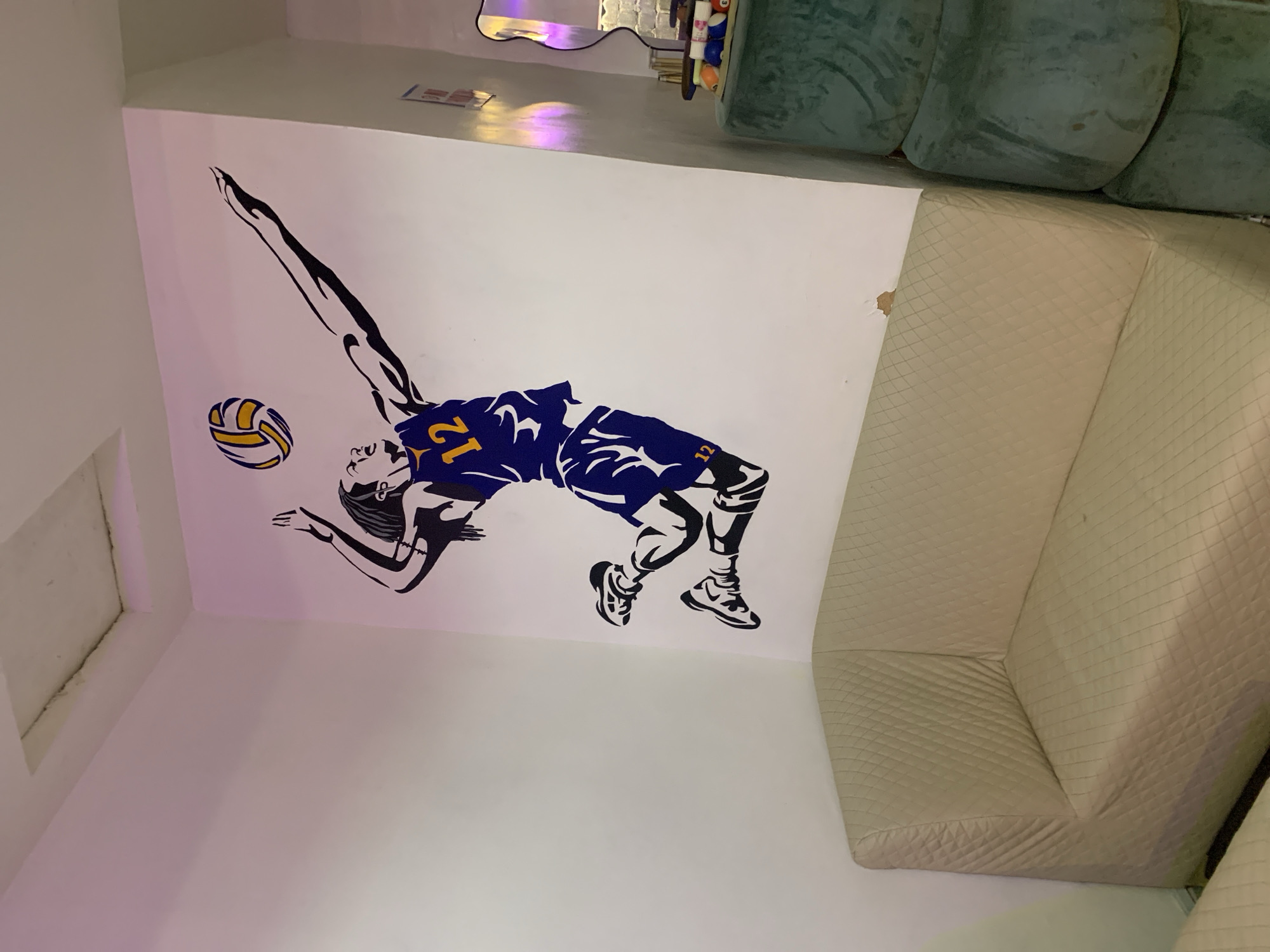 The image depicts an indoor scene with a volleyball-themed wall decal. The decal shows a stylized representation of a volleyball player diving to hit a volleyball. The player is wearing a jersey with the number 12. A beige quilted couch is positioned adjacent to the wall with the decal. A green couch is also visible above the quilted one. There is an air vent on the wall to the left of the decal. The scene is likely in a private residence or recreational space. There is no information provided to determine if the location is in the city of Barakin Akawo, Nigeria.