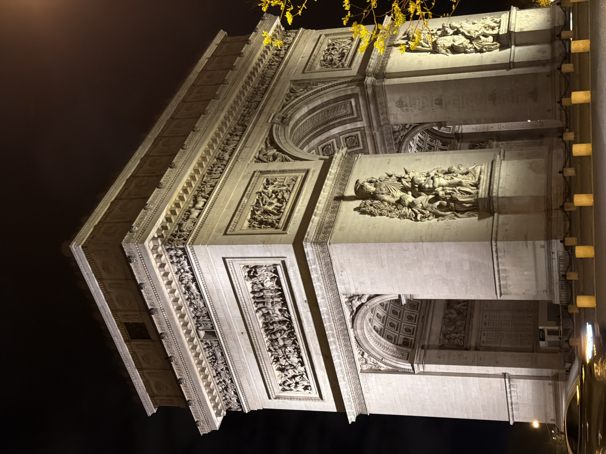 This image depicts the Arc de Triomphe in Paris, France at night. The monument is illuminated, highlighting its intricate carvings and architectural details. The sky is dark, emphasizing the bright lighting on the structure. There are decorative lights to the right of the image. There are yellow flowers on top of the building, possibly from nearby foliage. The perspective is from a low angle, looking up at the arch.