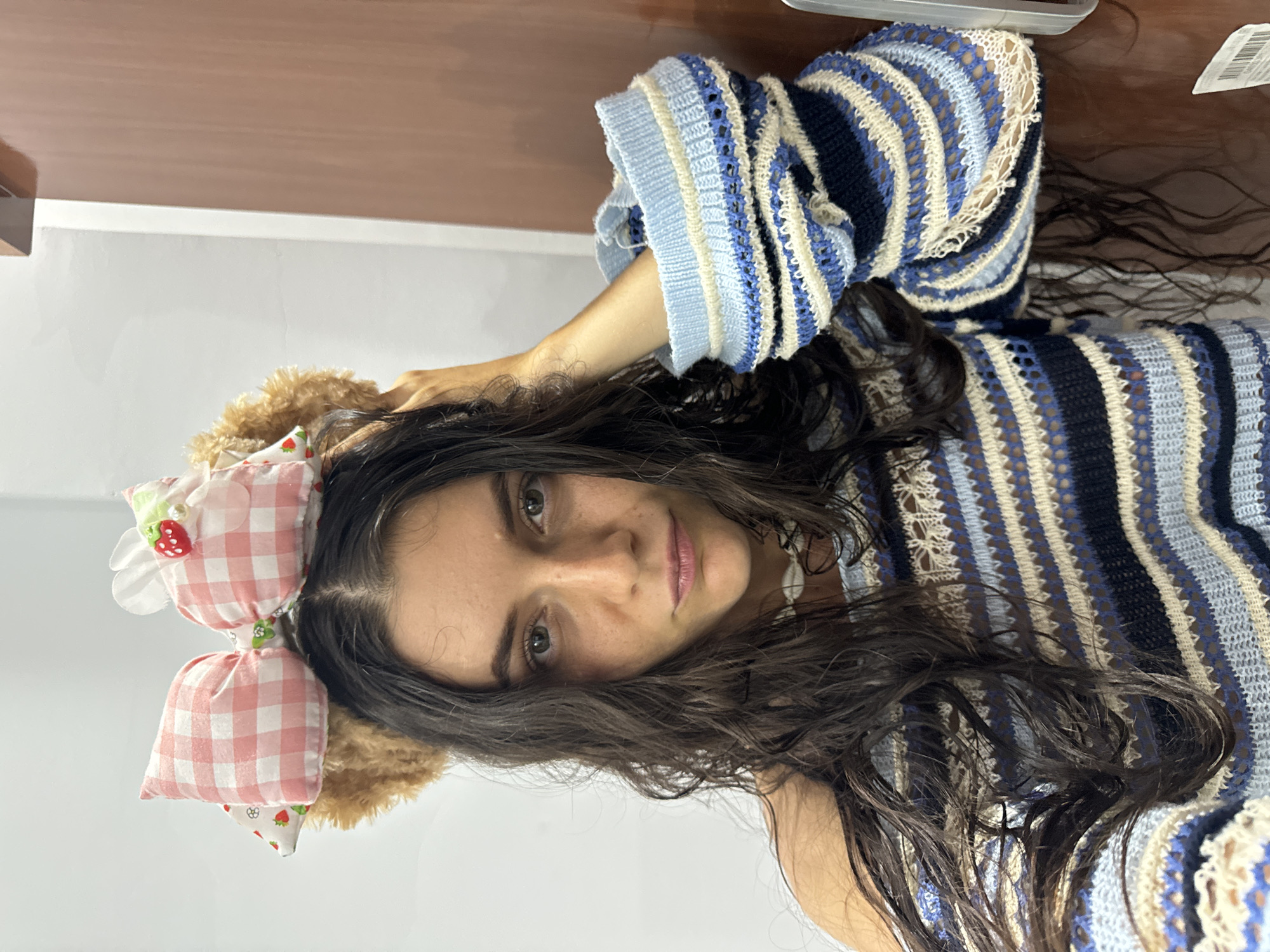 A portrait-style image features a young woman with long, dark, wavy hair looking directly at the camera. She is wearing a distinctive knit sweater with horizontal stripes in shades of dark blue, light blue, and cream, characterized by an open, airy pattern. On her head, she wears a fluffy, light brown headband from which two prominent pink and white gingham bows extend. One of these bows is adorned with a small, red strawberry embellishment and a white flower detail.
Her face is framed by her dark hair, revealing light-colored eyes, a straight nose with visible freckles, and light pink lips. Her expression appears calm and neutral. Her right hand is raised, with fingers gently touching her head near the headband, suggesting a casual pose or an adjustment of her accessory.
The background consists of a plain white wall, topped by a smooth, brown surface that resembles wood. The lighting is soft and even, indicating an indoor setting. There are no visual cues within the image to suggest the time of day or outdoor weather. While the provided context places the location as Mũi Né, Vietnam, this is not evident from the visual information alone. No text is visible in the media file.